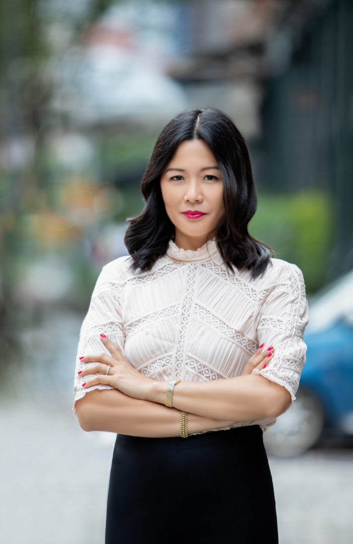 A woman with black hair wearing a white lace blouse and a black skirt standing outdoors with her arms crossed.