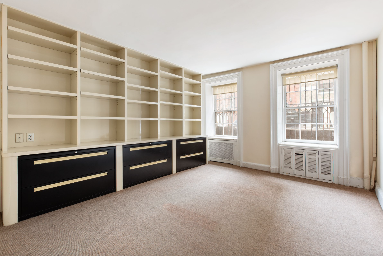 Empty room with beige carpet, built-in shelving unit with black drawers, and two large windows with white trim and vertical blinds, allowing sunlight to enter.