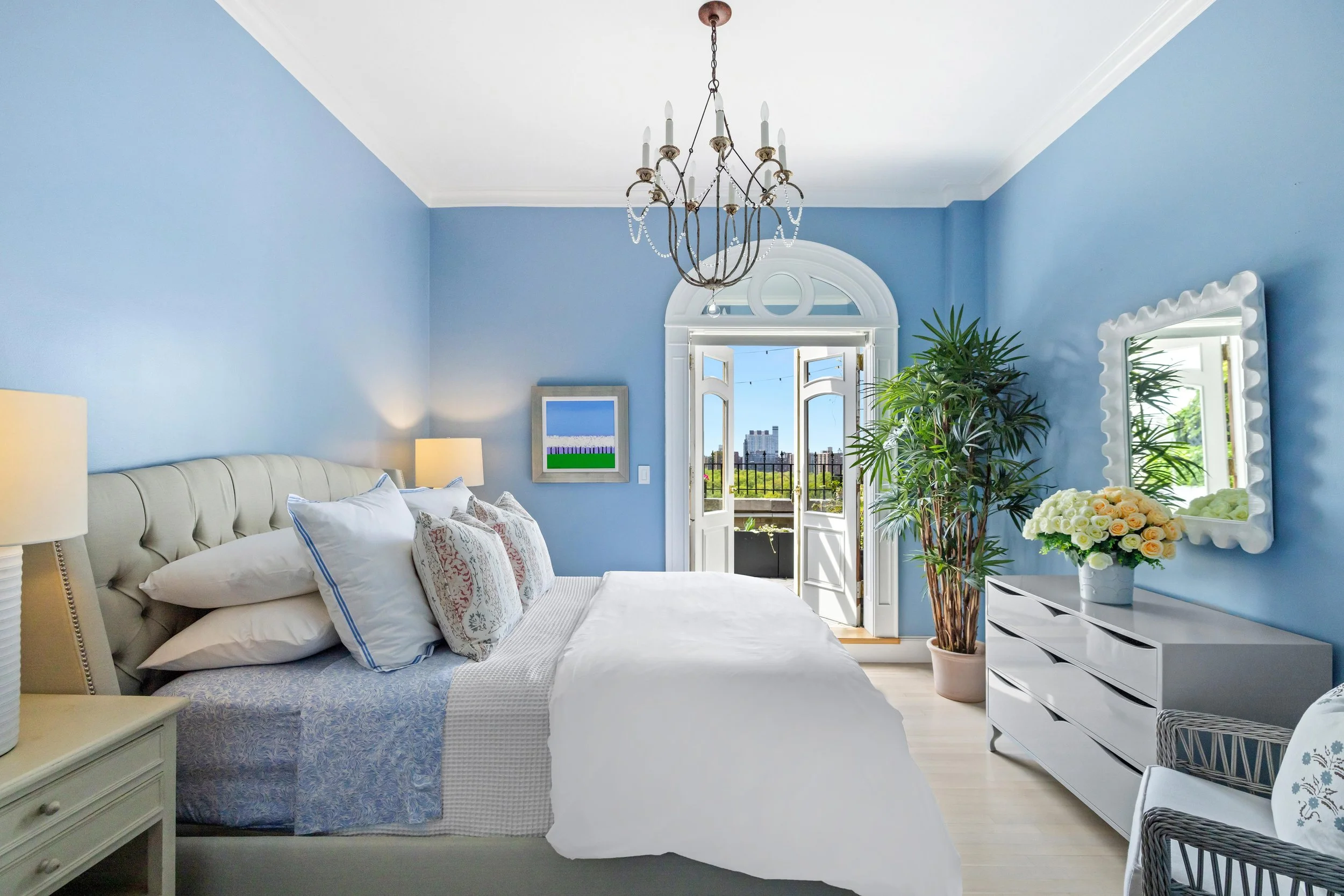 Bright bedroom with blue walls, white bed, and a balcony door with a city view, decorated with a hanging chandelier, potted plant, and mirror.