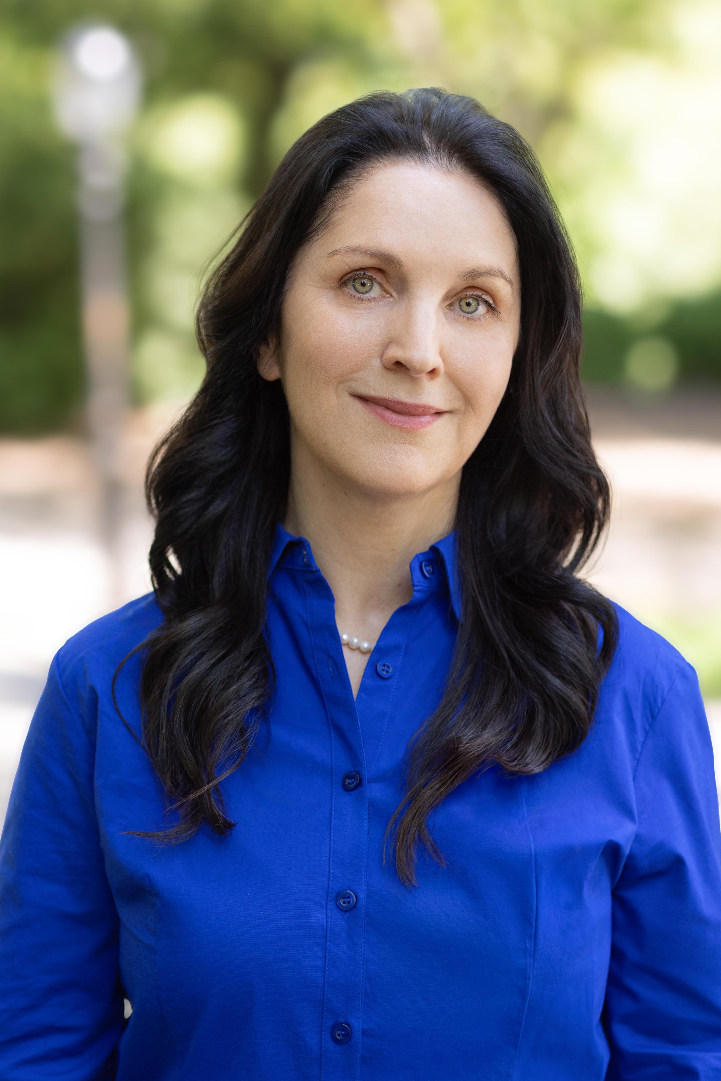 A woman with long dark hair, light skin, and light eyes, smiling and wearing a blue button-up shirt and pearl necklace, outdoors with blurred green trees in the background.