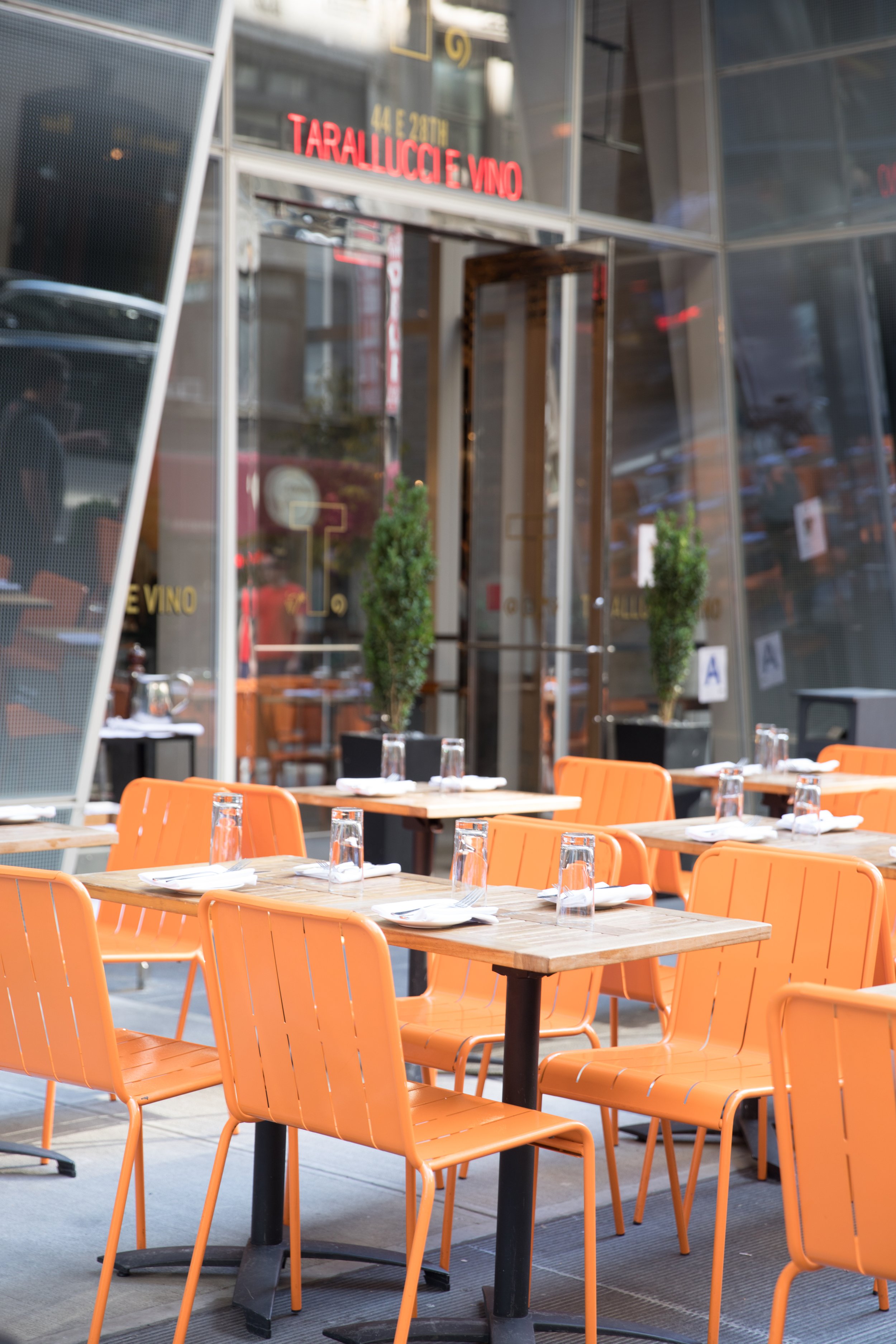 Empty restaurant patio with orange chairs, wooden tables, upside-down glasses, and utensils set on the tables outside of a restaurant with a glass facade and green plants.