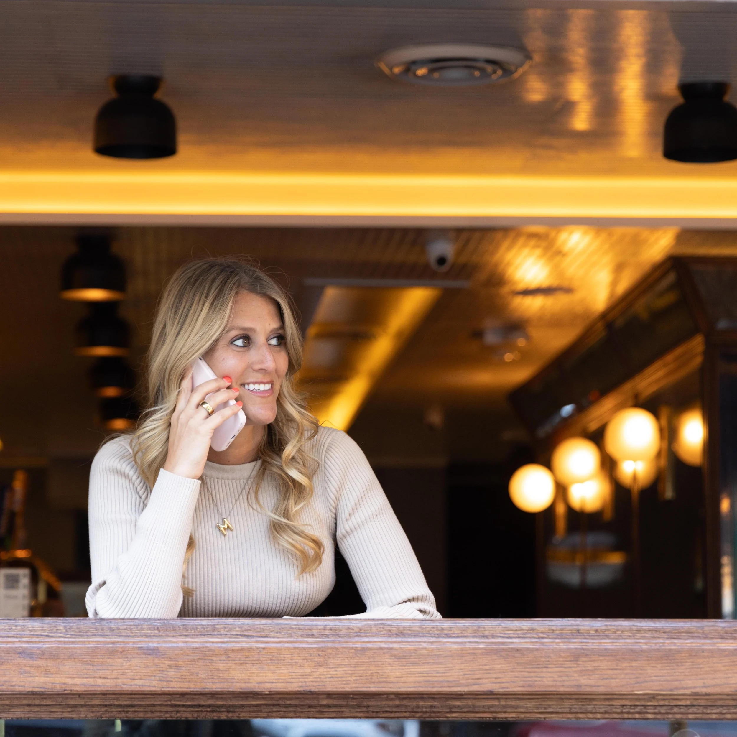 A woman sitting at a bar or table, talking on a mobile phone, in a warmly lit indoor setting.