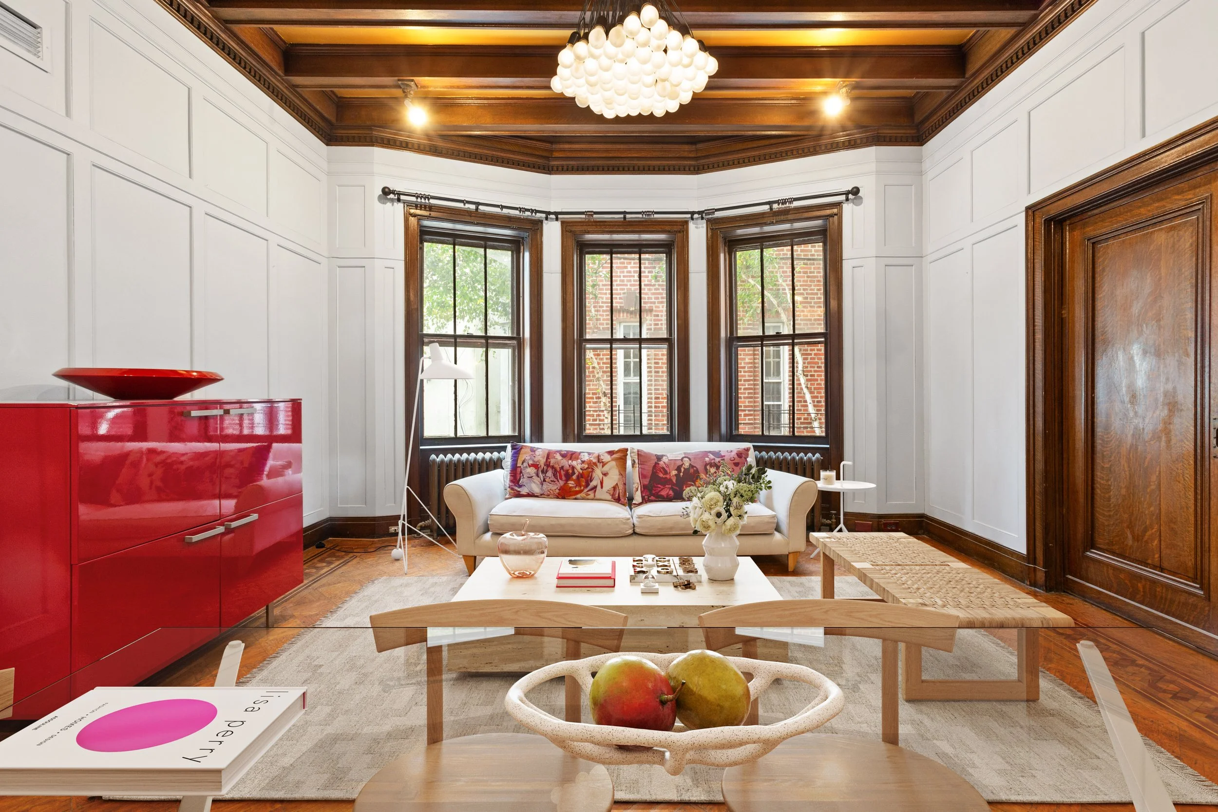 Living room with white walls, wooden ceiling, and large bay windows. Features a white sofa with colorful pillows, a red sideboard, a coffee table with books and flowers, and a bowl of fruit in the foreground.