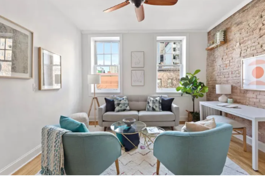 Bright living room with white walls, brick accent wall, large windows, and modern furniture including a beige sofa, teal armchairs, a glass coffee table, and decorative pillows. Artwork and a potted plant are also visible.