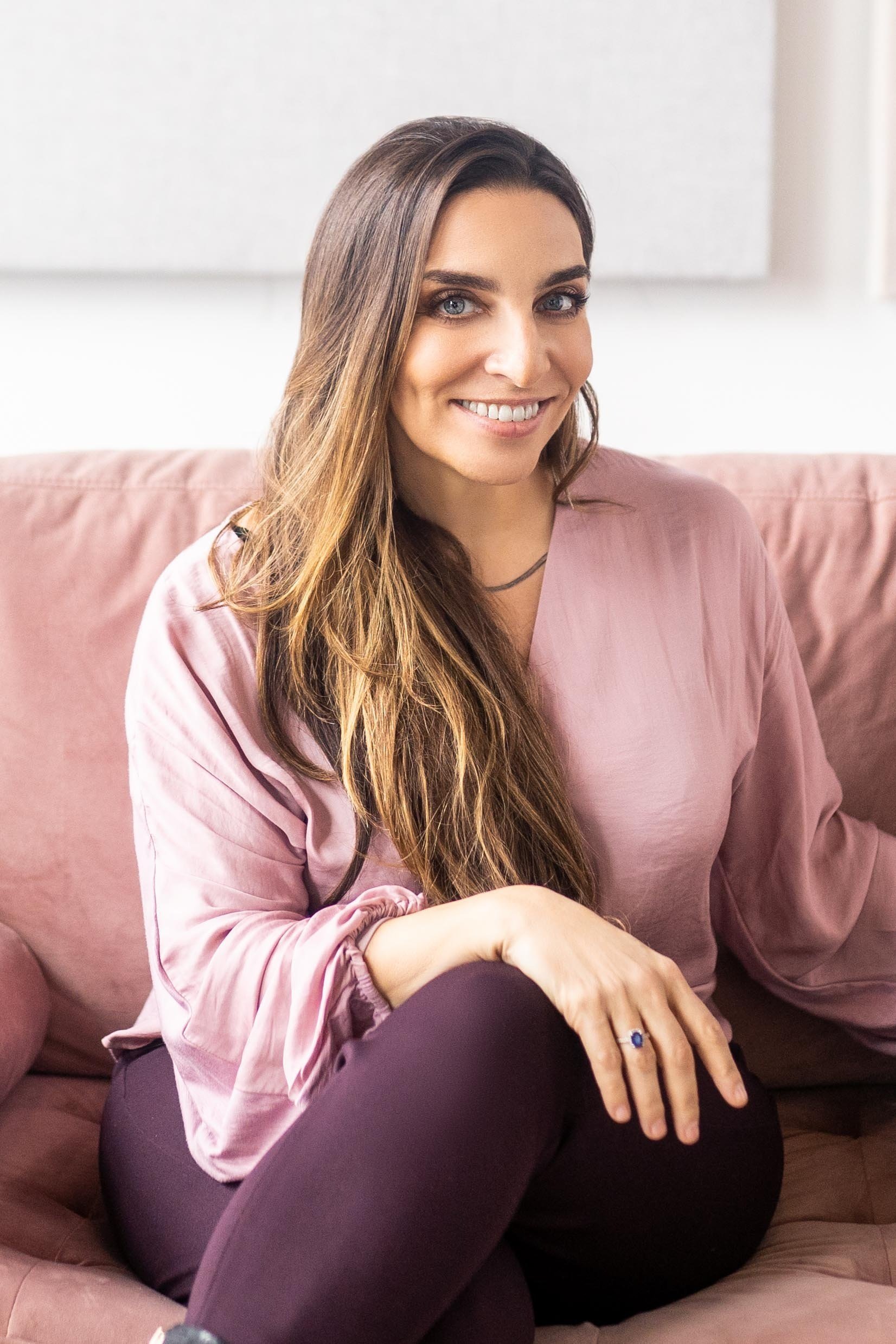 A woman with long brown hair and blue eyes sitting on a pink couch, smiling at the camera.