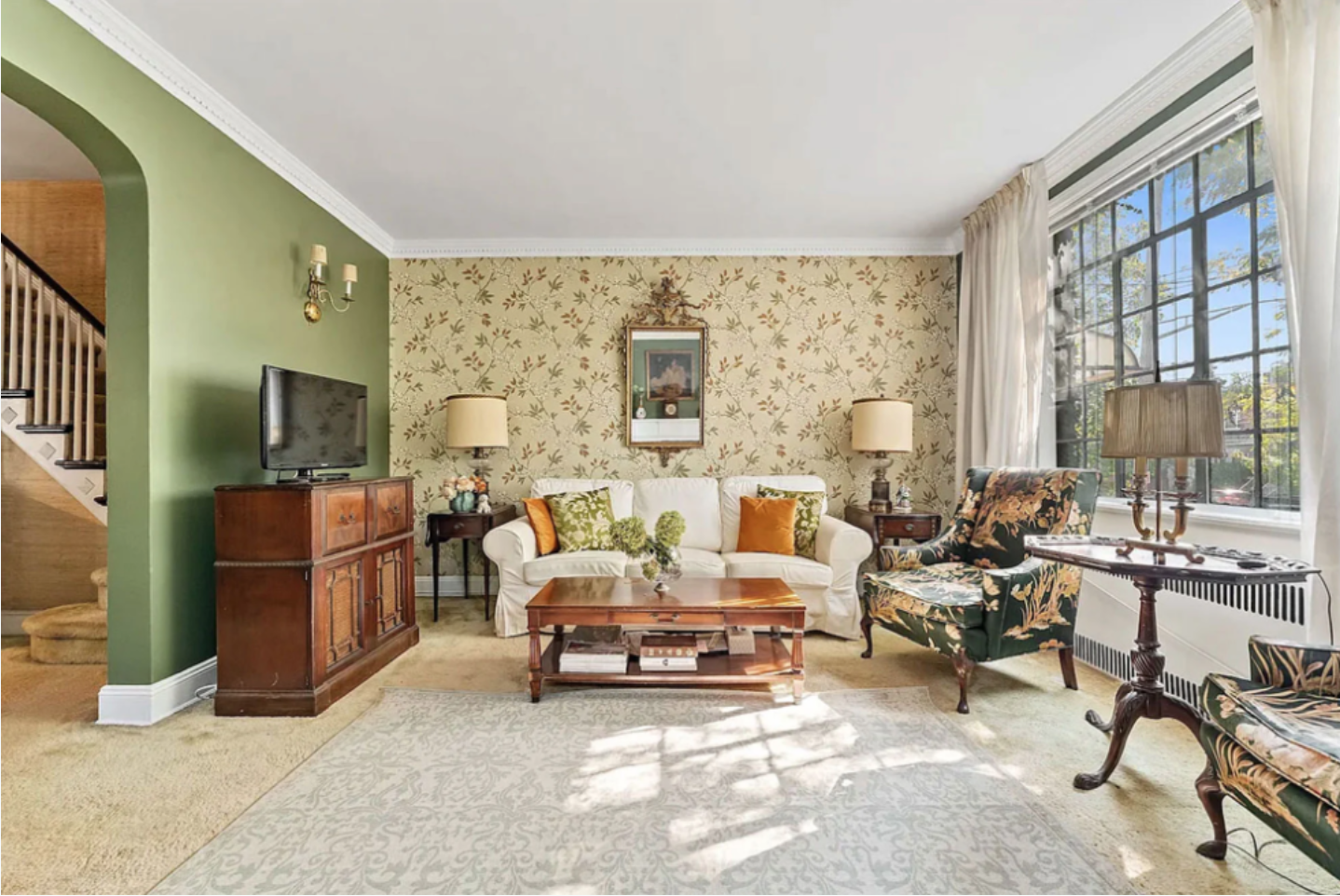 Living room with green wall on the left, floral wallpaper on the far wall, a white sofa with orange and green pillows, a wooden coffee table, floral armchairs, a window with white curtains, and a big flat-screen TV on a wooden stand.