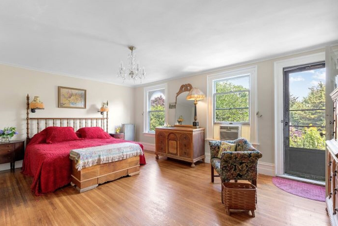 A cozy bedroom with a large bed covered in a red blanket and matching pillows, wooden furniture including a dresser with a mirror, a nightstand, a small armchair with floral fabric, a wicker basket, and a door leading to a balcony with trees visible outside.