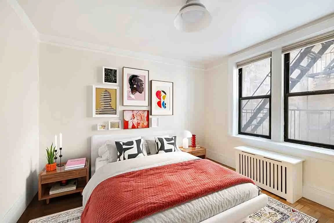 A bright bedroom with a bed covered in a coral blanket and black and white pillows, wooden nightstands with decorative items, framed artwork on the wall, large windows with black frames, and hardwood floors.