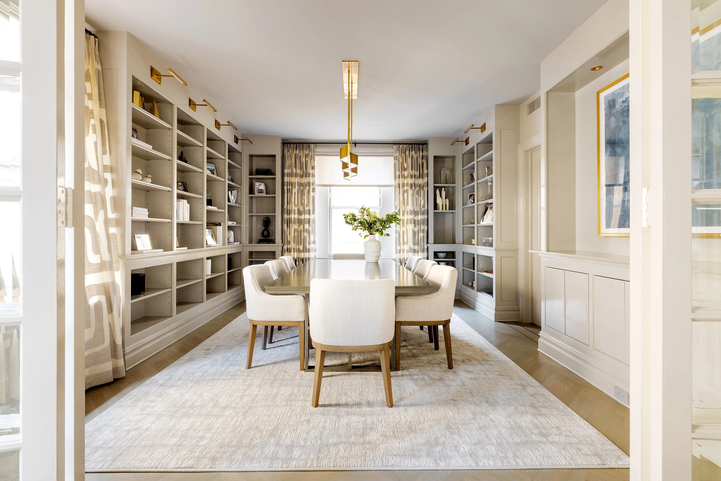 Bright dining room with a wooden table, six upholstered chairs, built-in shelves filled with books and decorative items, a large window with patterned curtains, and a modern gold chandelier.