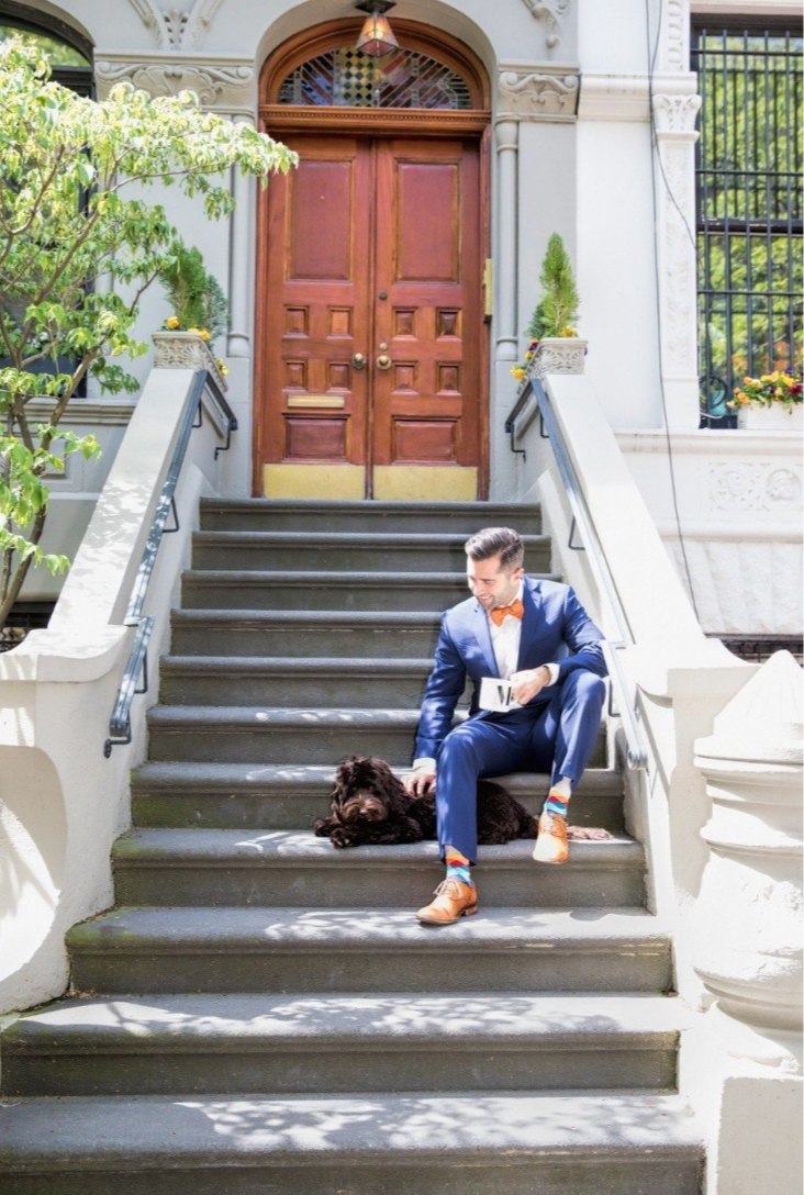 A man in a blue suit with colorful socks and brown shoes sitting on outdoor stairs, holding a document, with a black dog lying beside him. The stairs lead up to a large wooden door with potted plants on the sides, and a building with ornate architect