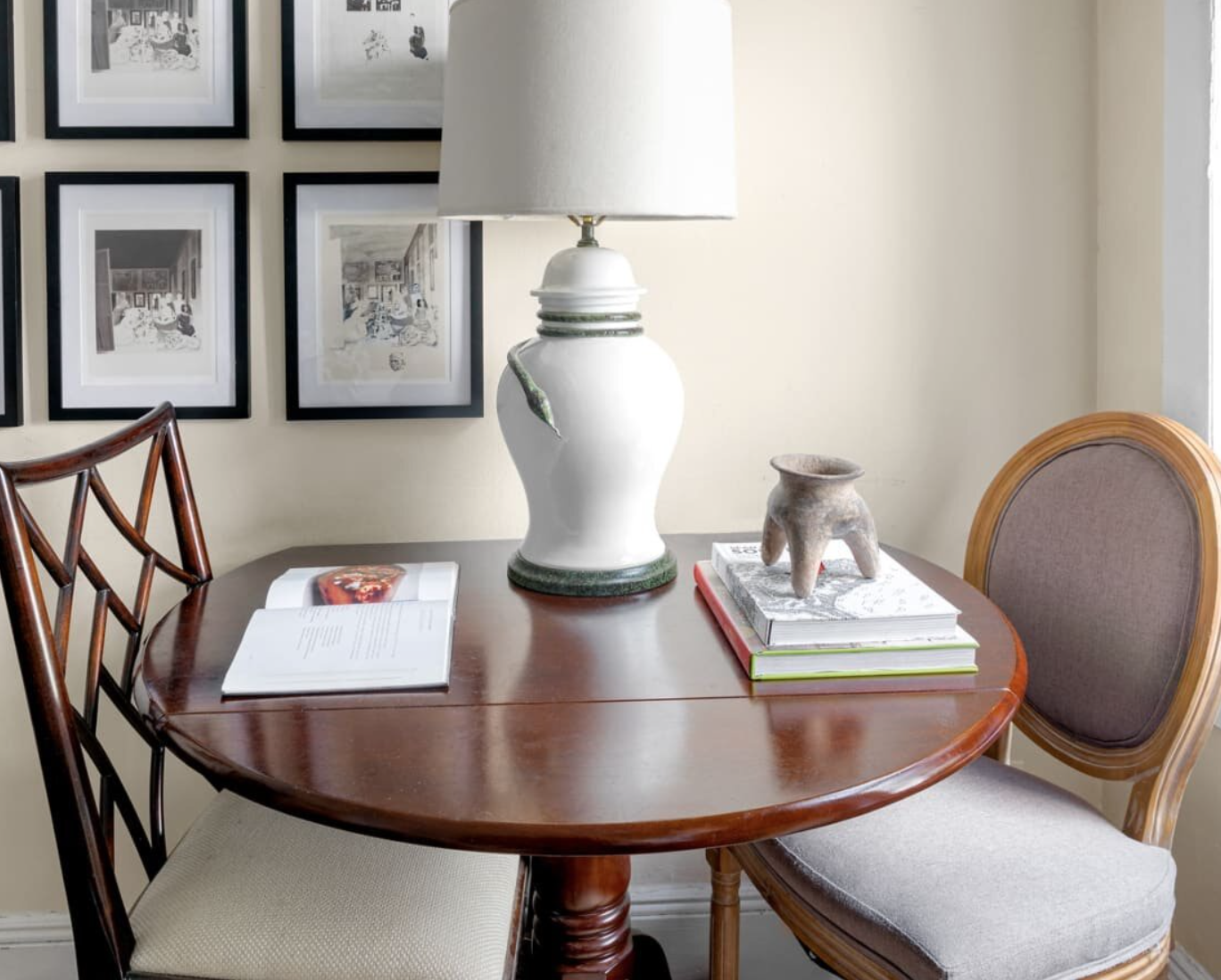 A wooden dining table with a large white ceramic table lamp, books, and a small pottery sculpture. There are two chairs, one with a lattice backrest and the other with a rounded wooden frame and upholstered seat. Behind the table, a wall displays a collection of framed black and white photographs.
