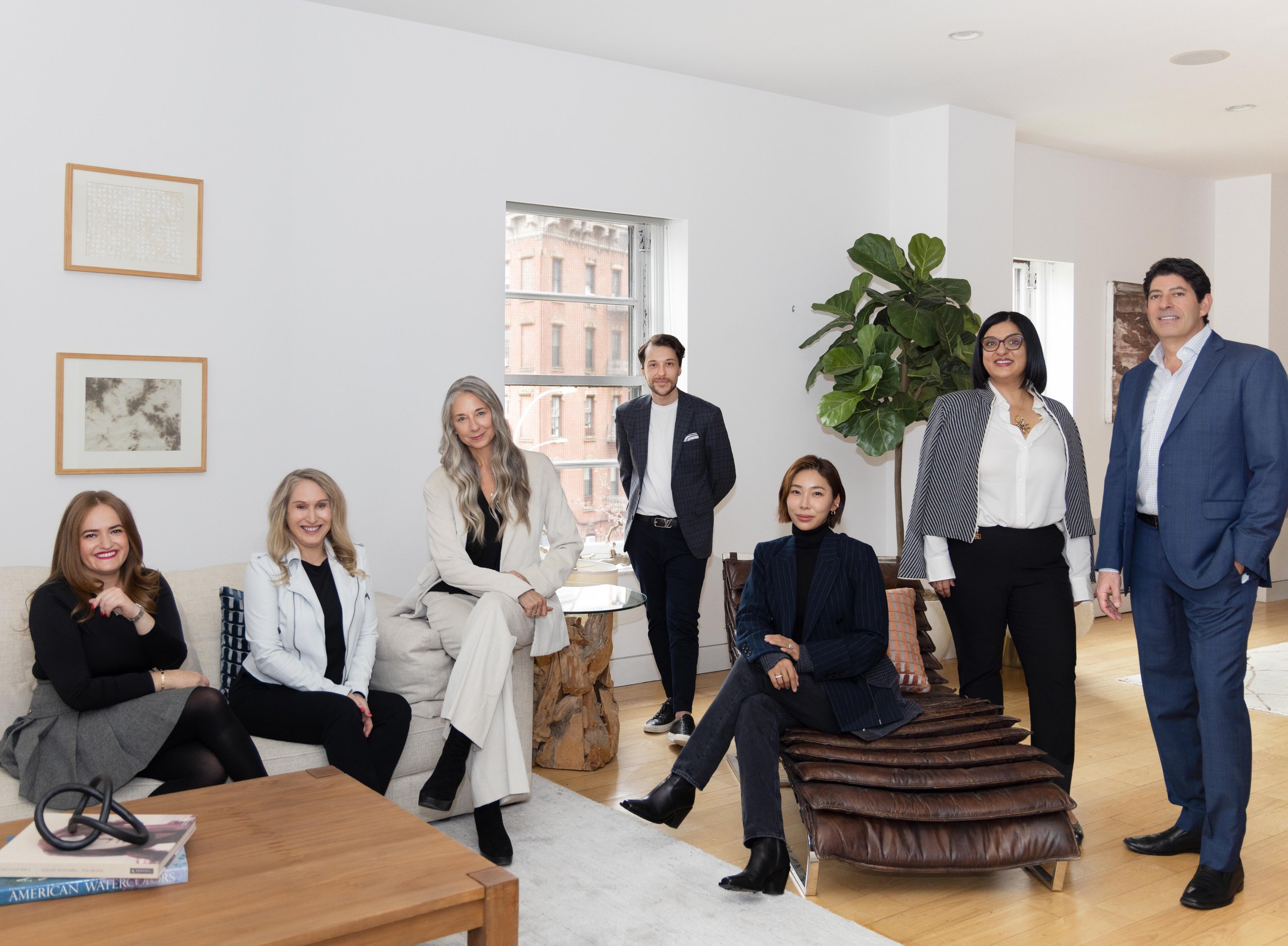 Group of seven diverse professionals in a modern office lounge, smiling and posing for a photo, with a large window and a green plant in the background.