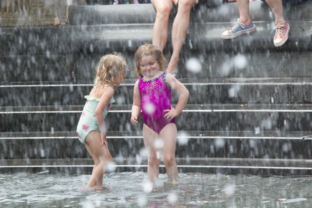 Two young girls in swimsuits playing in a fountain splash area, with water falling around them, while adults sit on the steps above.