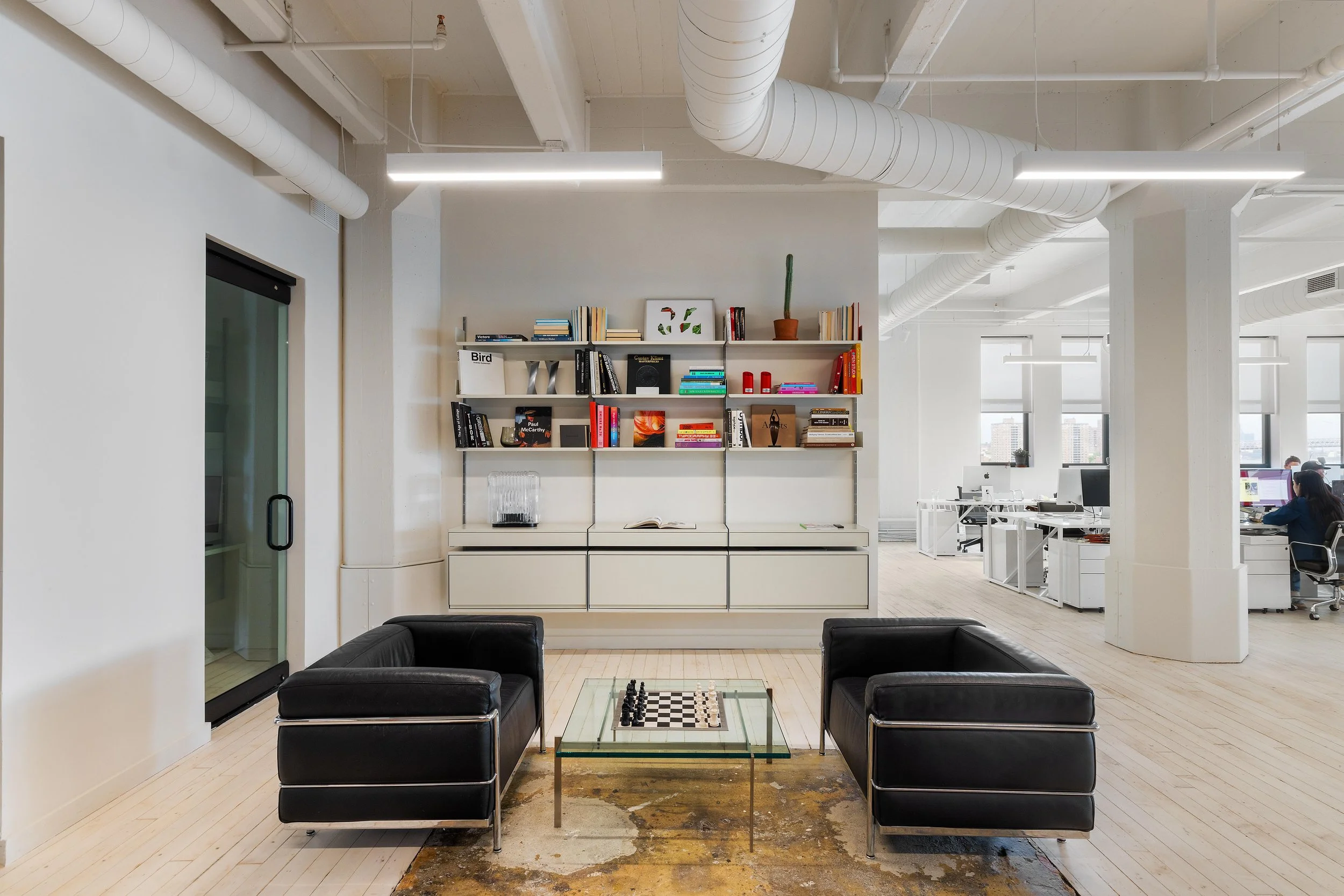 Modern office lounge area with two black leather sofas facing each other, a glass chess table in between, white built-in shelving with books and decor, and open office space with desks and employees in the background.
