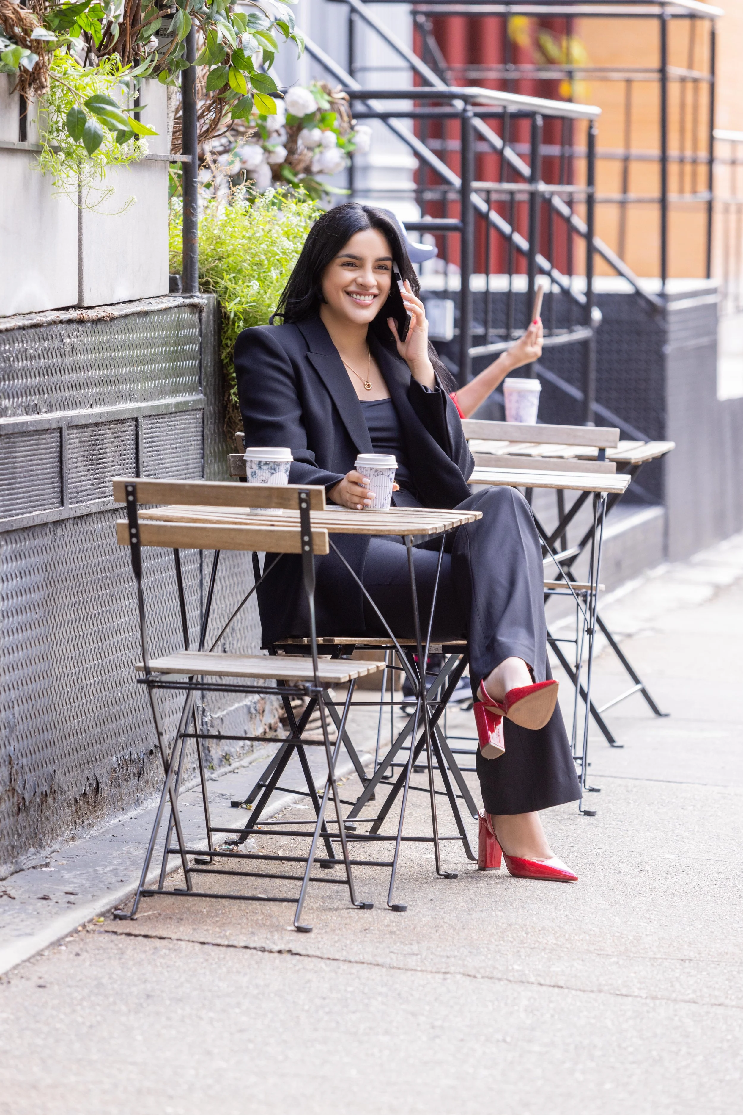 A woman sitting outside at a cafe table, smiling while talking on her cell phone. She is wearing a black blazer, black pants, and red high heels. There are coffee cups on her table, and the sidewalk and railing are visible in the background.