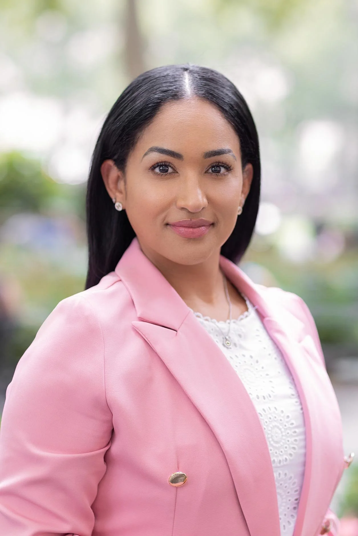 A woman with long black hair, wearing a light pink blazer, white lace top, pearl earrings, and a necklace, stands outdoors with a blurred background of trees and lights.