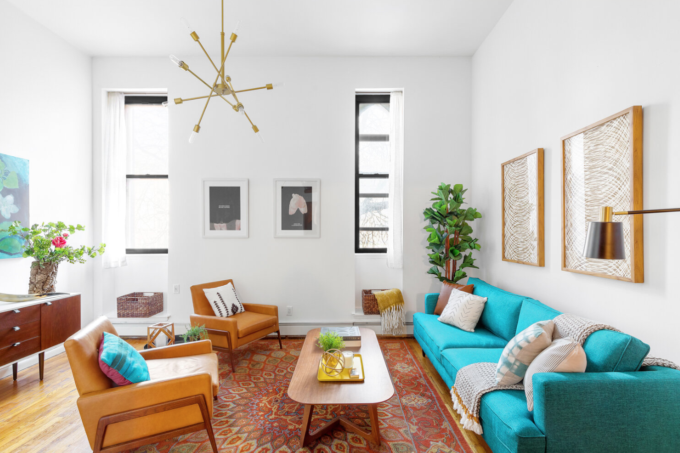 Bright living room with white walls, large windows with white curtains, teal and tan sofas, wooden coffee table with yellow tray and greenery, orange patterned rug, artwork on walls, green plant, and modern chandelier.