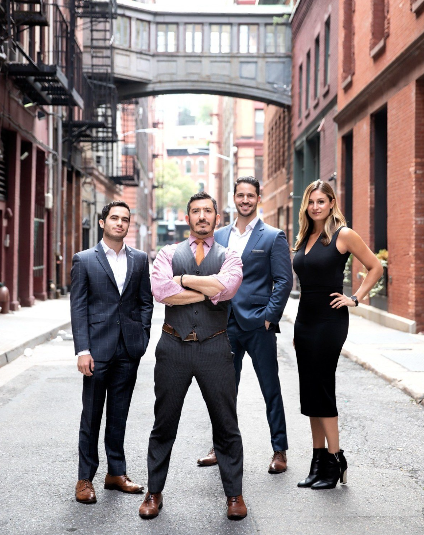 Group of four professionally dressed people standing in an urban alleyway with brick buildings and a pedestrian bridge overhead.
