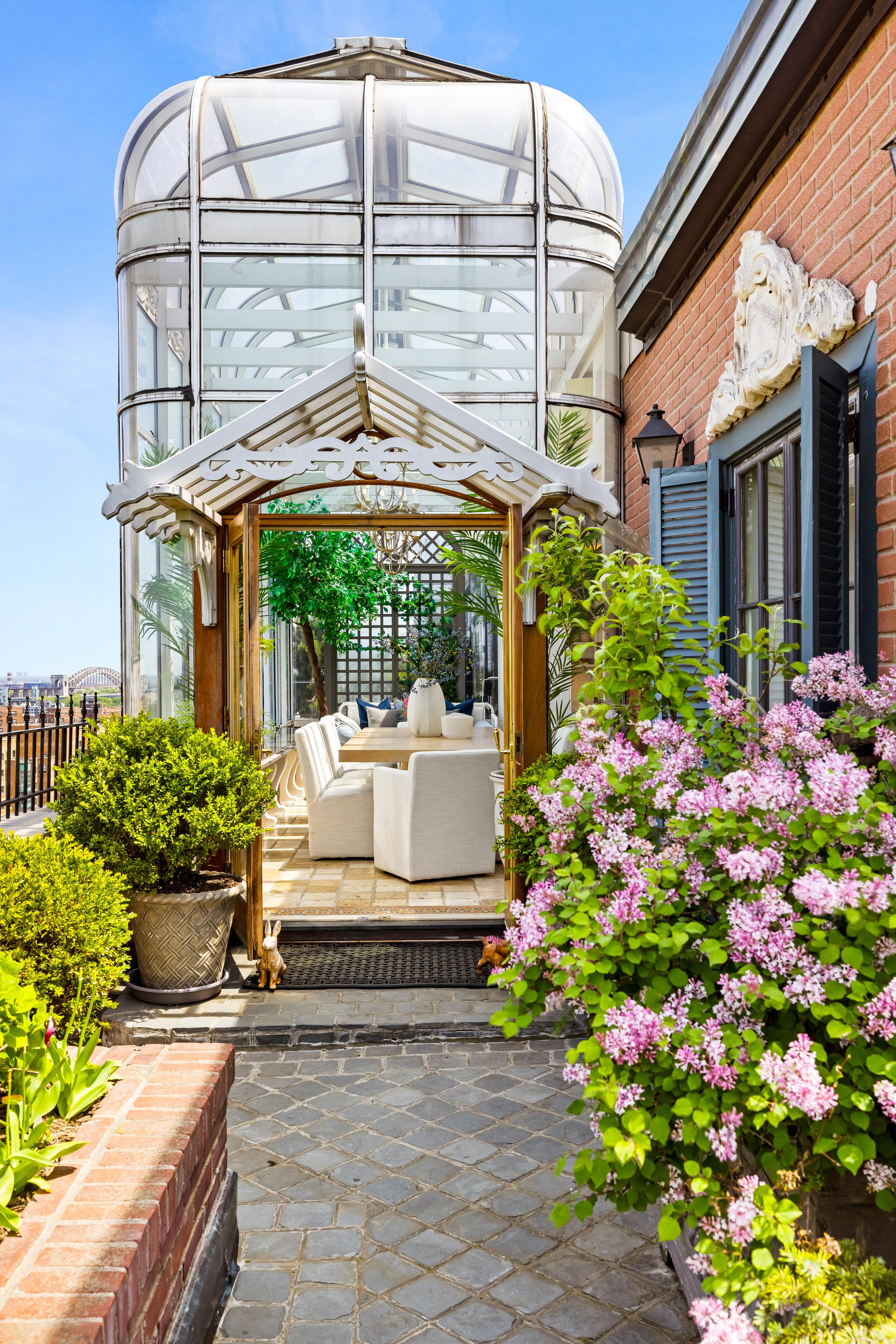 A cozy outdoor patio with lush greenery, pink flowers, and a glass greenhouse structure leading into a bright indoor seating area with white chairs and a wooden table.