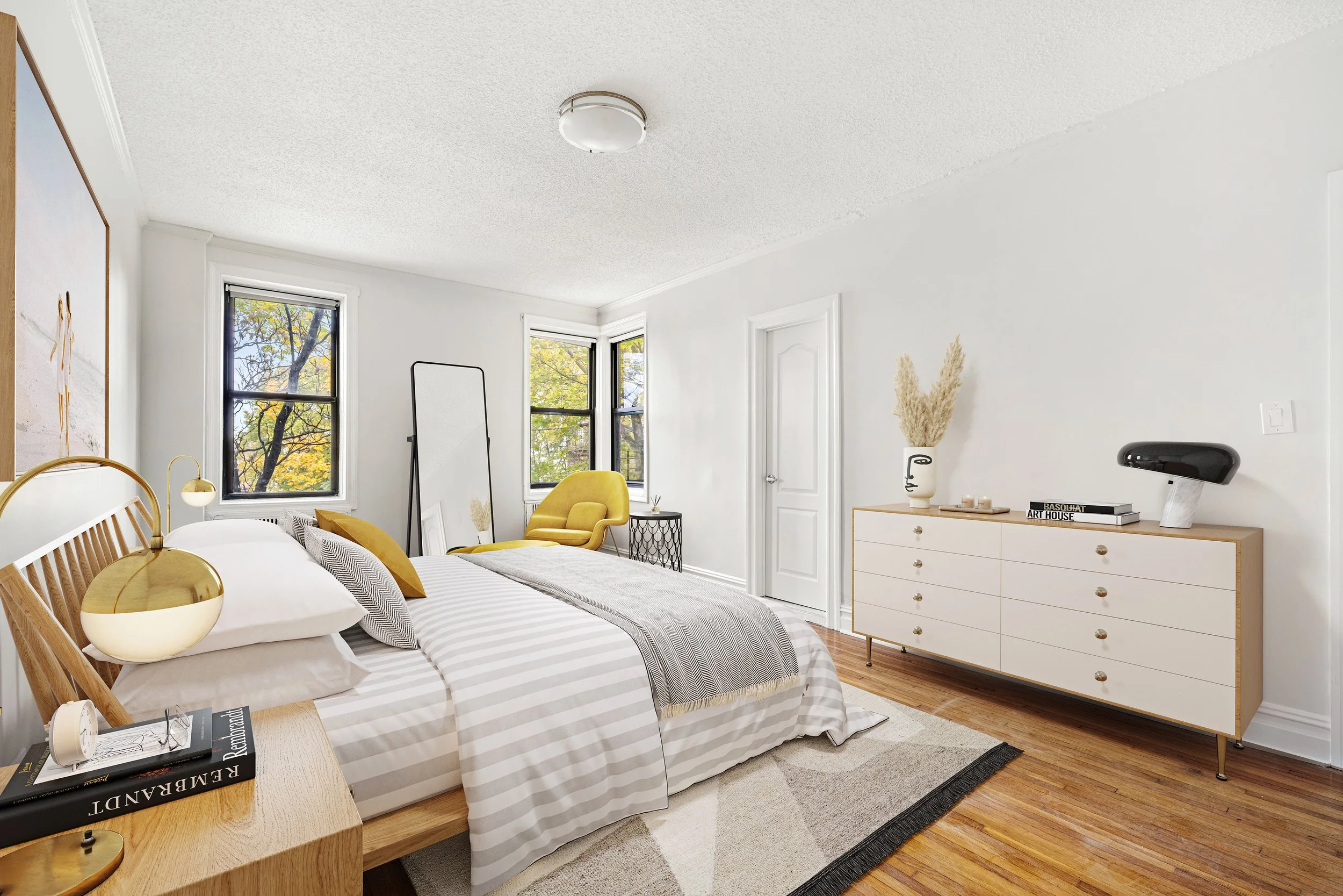 Bedroom with white walls, a bed with striped bedding, a yellow chair, a dresser with decor, and three windows showing trees outside.