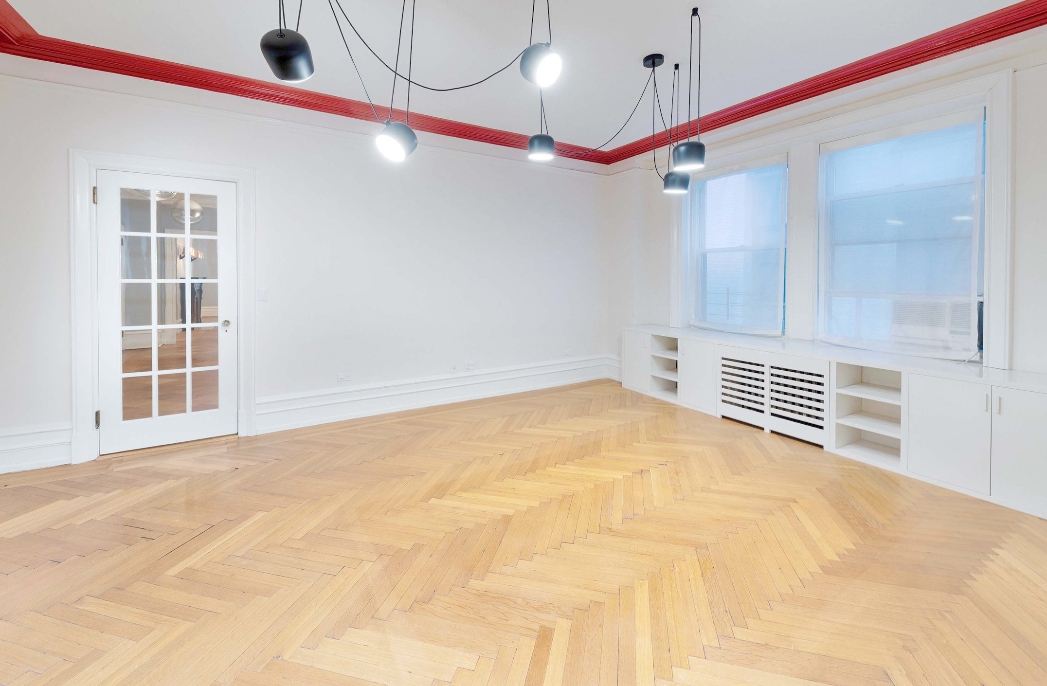 Empty room with wooden herringbone flooring, white walls, a door with glass panes, and large windows with blue shades. Modern ceiling lights hanging from black cords.