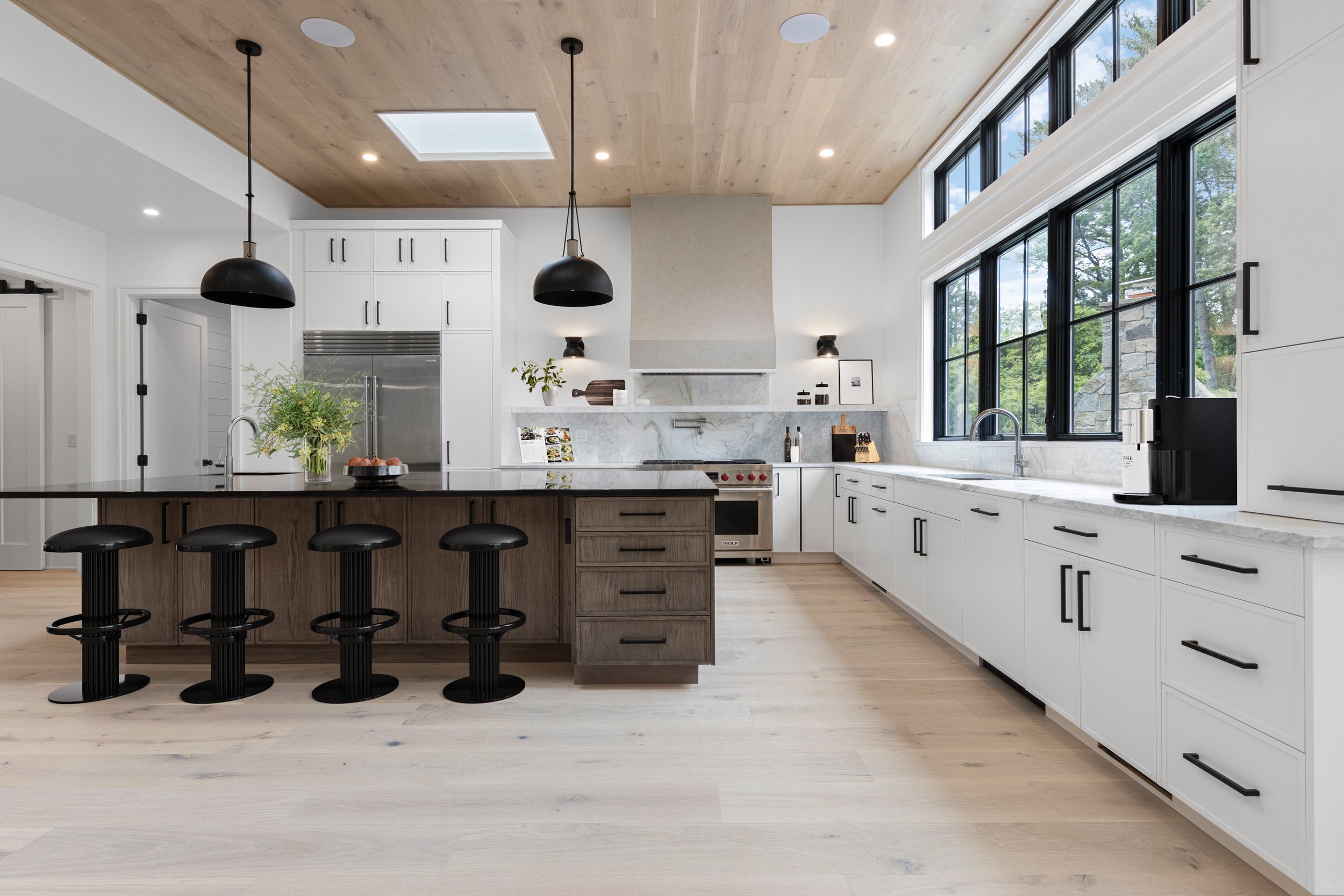 Modern kitchen with white cabinets, black hardware, large windows, a wooden ceiling, and a central island with black bar stools.