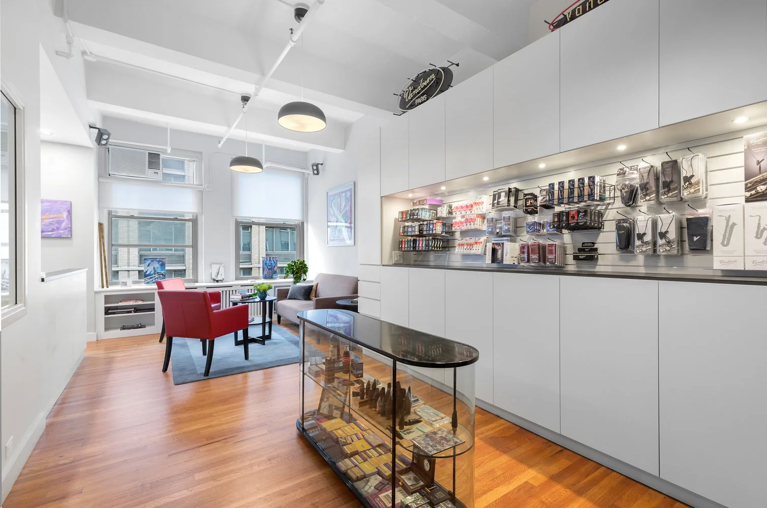 Inside a modern retail store with white walls and wooden floors, displaying various electronic accessories on wall-mounted shelves and clear display cases, a seating area with red and beige chairs by windows, and a small glass display table in the fo