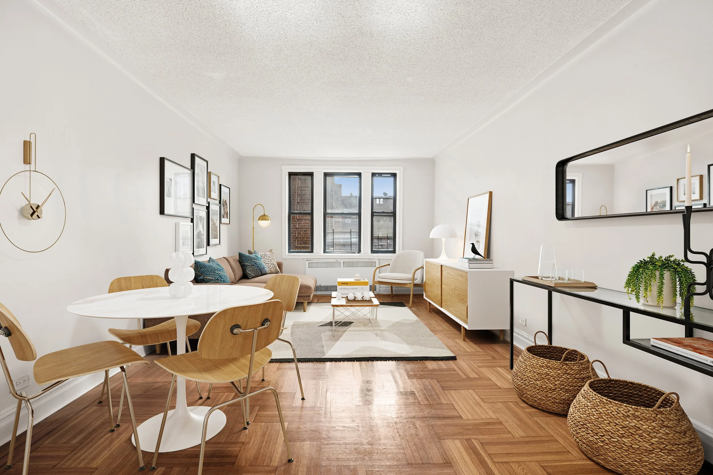Bright living room with white walls, wooden floor, and modern furniture including a round dining table with wooden chairs, a beige sofa with blue pillows, a white armchair, a wooden sideboard, and wall art. Large windows provide natural light.