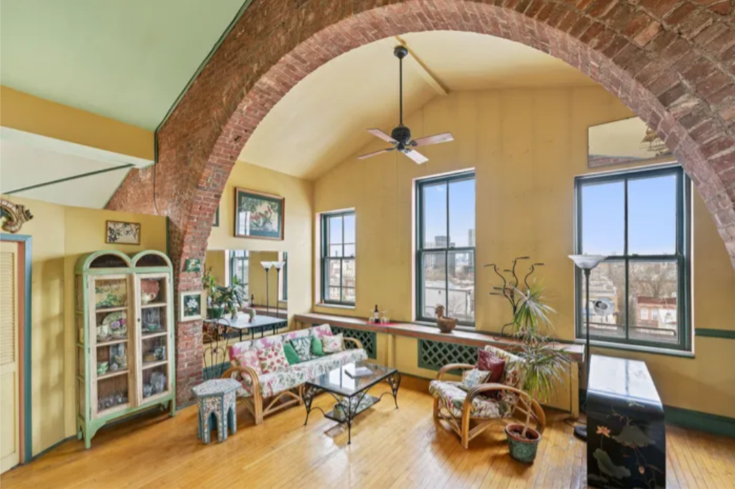 Living room with yellow walls, large arched brick ceiling, and natural light from three windows. Features a wicker floral-patterned sofa, matching armchair, glass coffee table, potted plants, a floor lamp, and decorative items.
