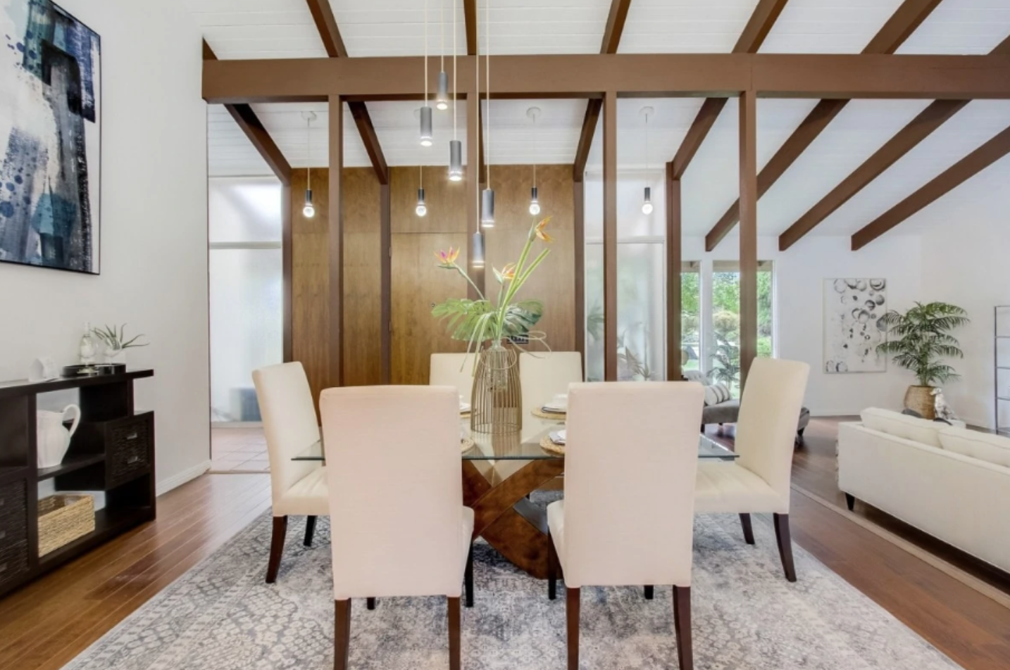 A dining room with a glass-top table and six cream-colored chairs. A vase of flowers is on the table. The room has hardwood floors, a patterned rug under the table, wooden ceiling beams, and modern hanging pendant lights. There is a wooden wall and large windows with outdoor greenery visible.