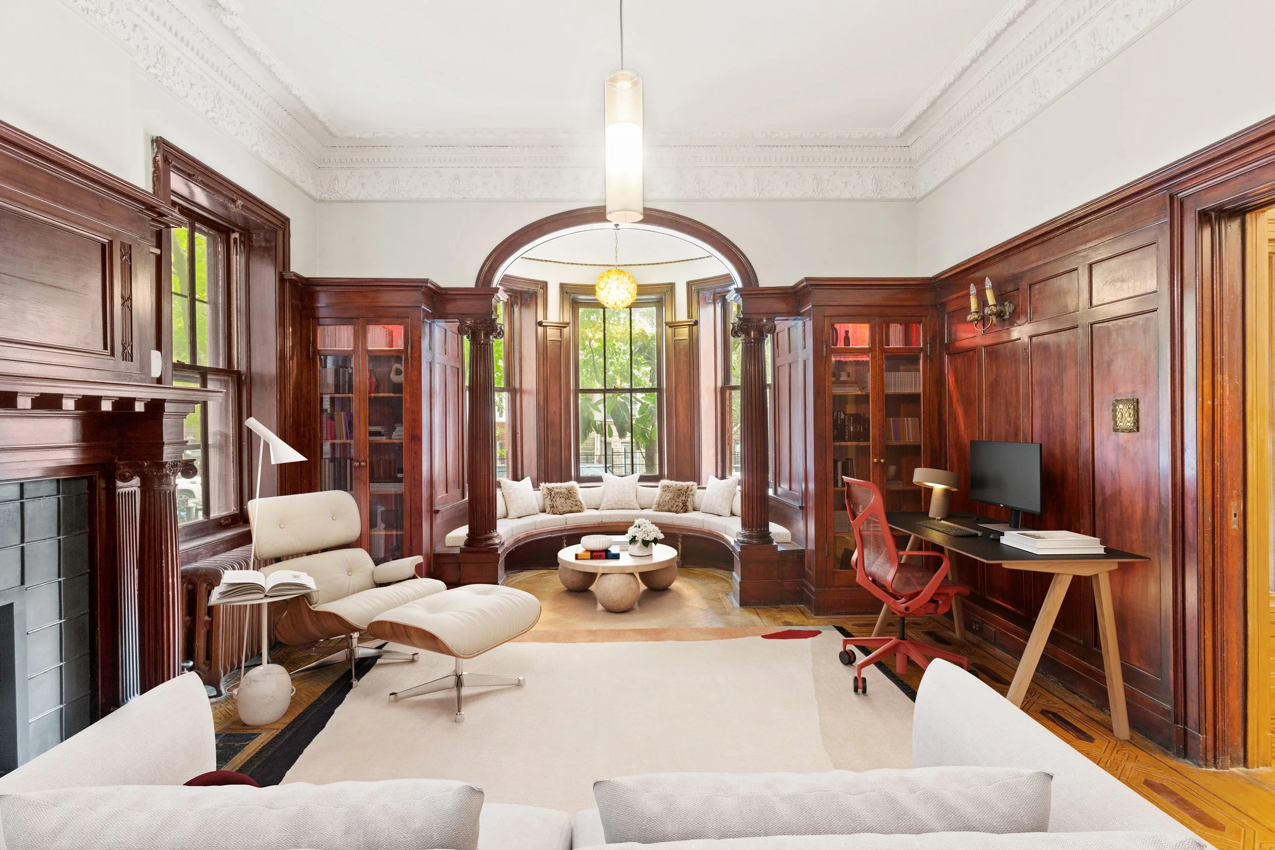 Living room with dark wood paneling, built-in shelves, large bay window with cushioned window seat, a white armchair with a book lamp, a red desk chair with a computer, and light-colored sofas.