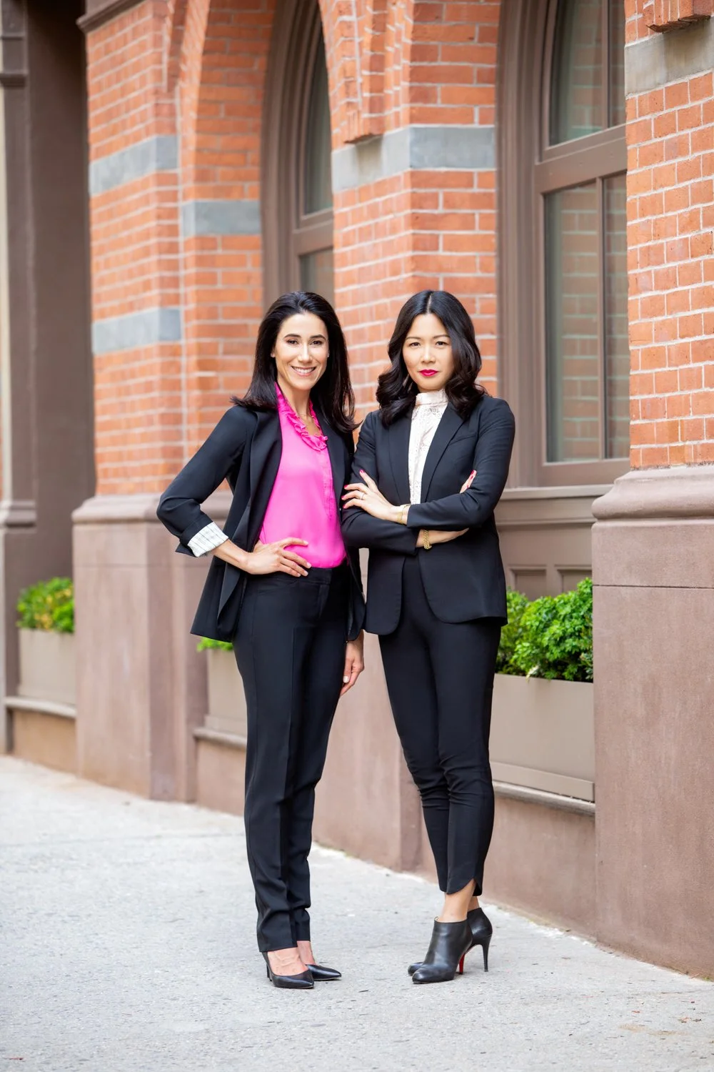 Two professional women in black suits, one with a pink blouse, standing outside a brick building with large windows.