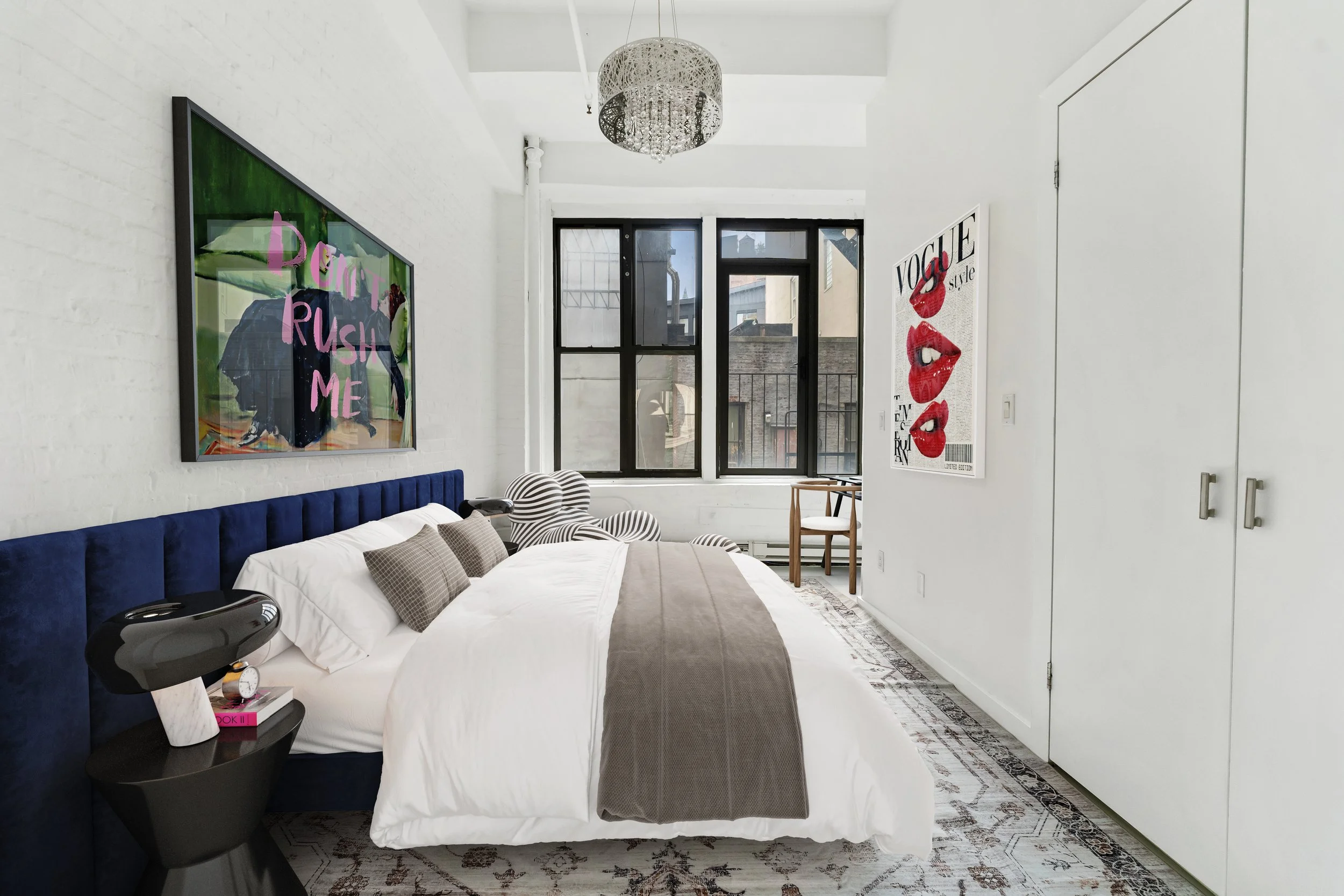 Modern bedroom with white walls, large black-framed windows, a blue upholstered headboard, white bedding with gray pillows and a taupe bed runner, a black nightstand with a black lamp, and colorful wall art posters. There's a chandelier hanging from 