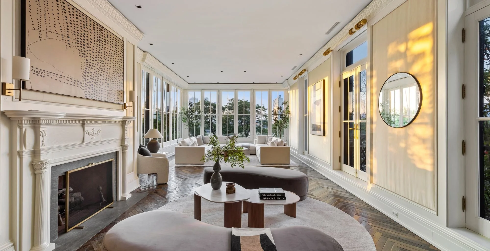 Bright living room with large windows, white sofas, a fireplace, and modern decor with natural light and trees visible outside.
