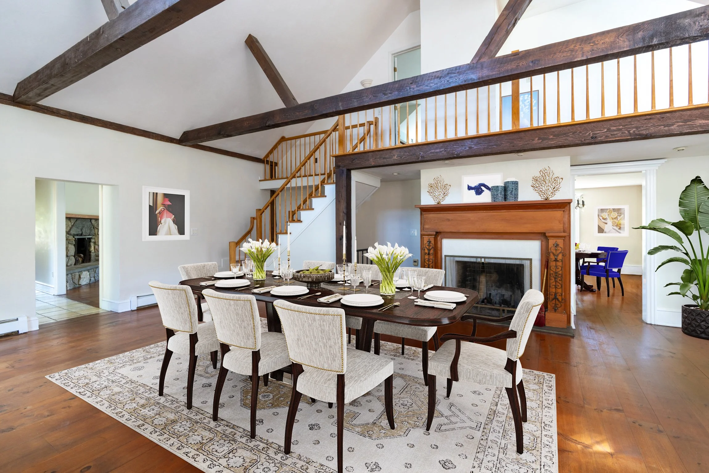 Dining room with a wooden table set for a meal, surrounded by upholstered chairs, a decorative fireplace, and a staircase leading to a second-floor balcony with wooden railing in a bright, spacious living area.