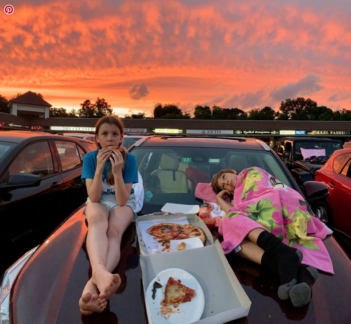 Two children relaxing on the hood of a parked car at sunset, with pizza and snacks, one sitting and the other lying down wrapped in a colorful blanket.