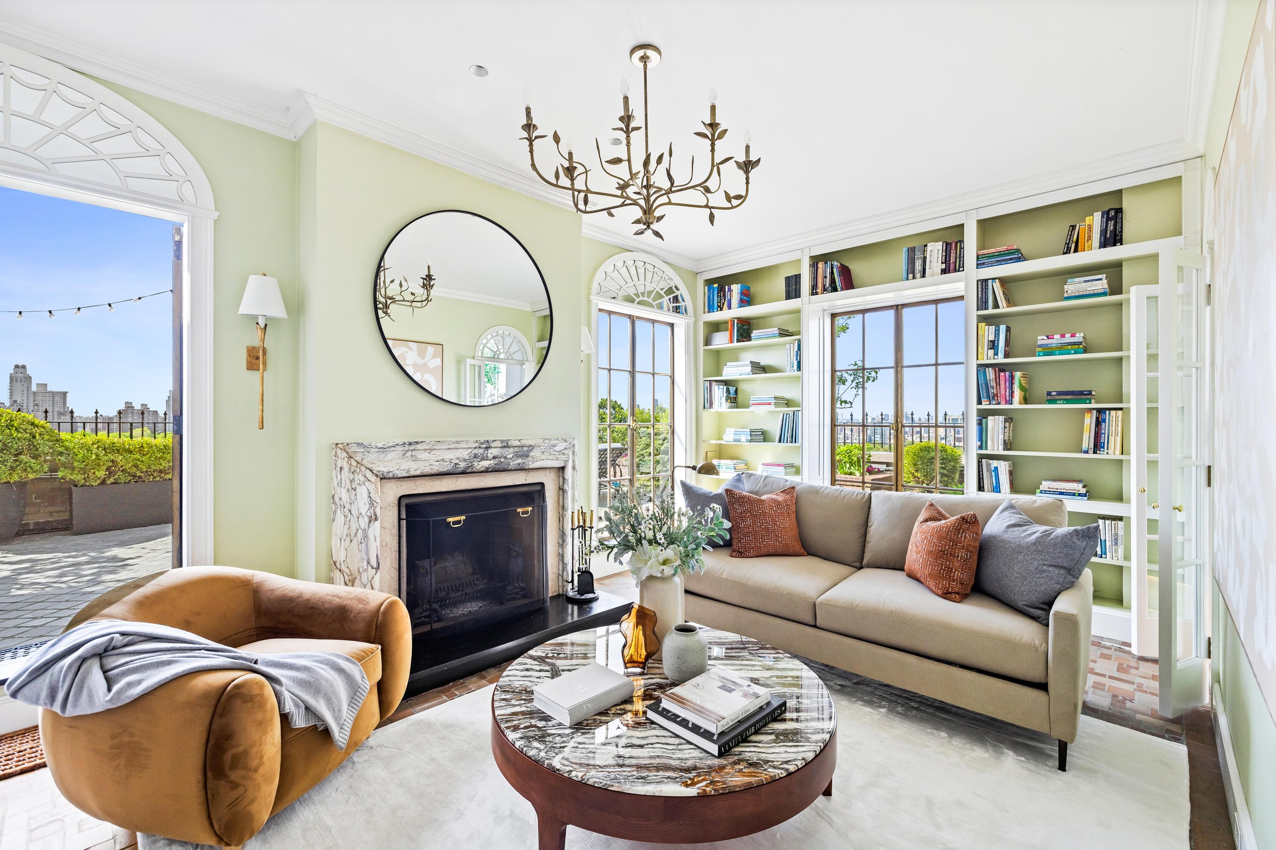 Bright living room with a beige sofa, an orange armchair with a gray blanket, a round marble-top coffee table, a marble fireplace, a wall mirror, built-in bookshelves filled with books, large windows showing a balcony with city views, and a chandelie