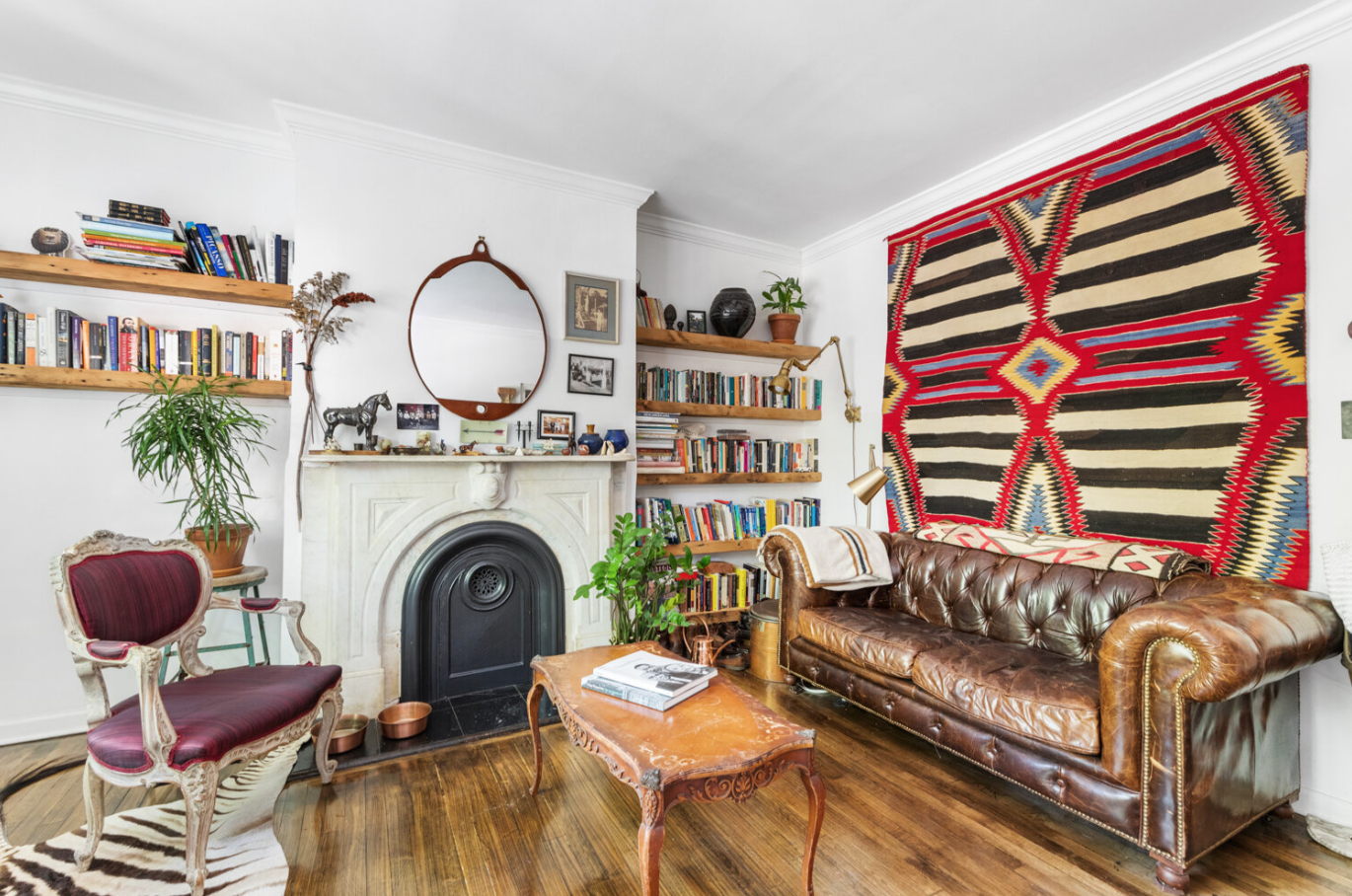 Living room with white walls, wooden bookshelves filled with books, a large colorful rug hanging on the wall, a leather sofa with a blanket, a vintage wooden coffee table, an ornate upholstered armchair, a mirror above a white fireplace, plants, and various decorative items.