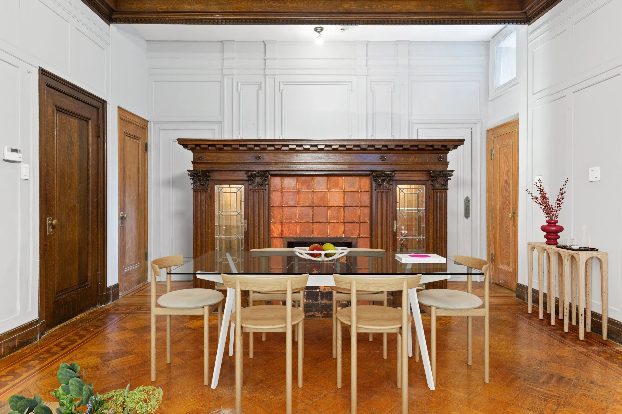 A dining room with a black glass-top table and six light wood chairs. In the background, a large ornate wooden fireplace with stained glass doors and a copper tile interior. To the right, a small side table with a red vase of dried flowers. The floor