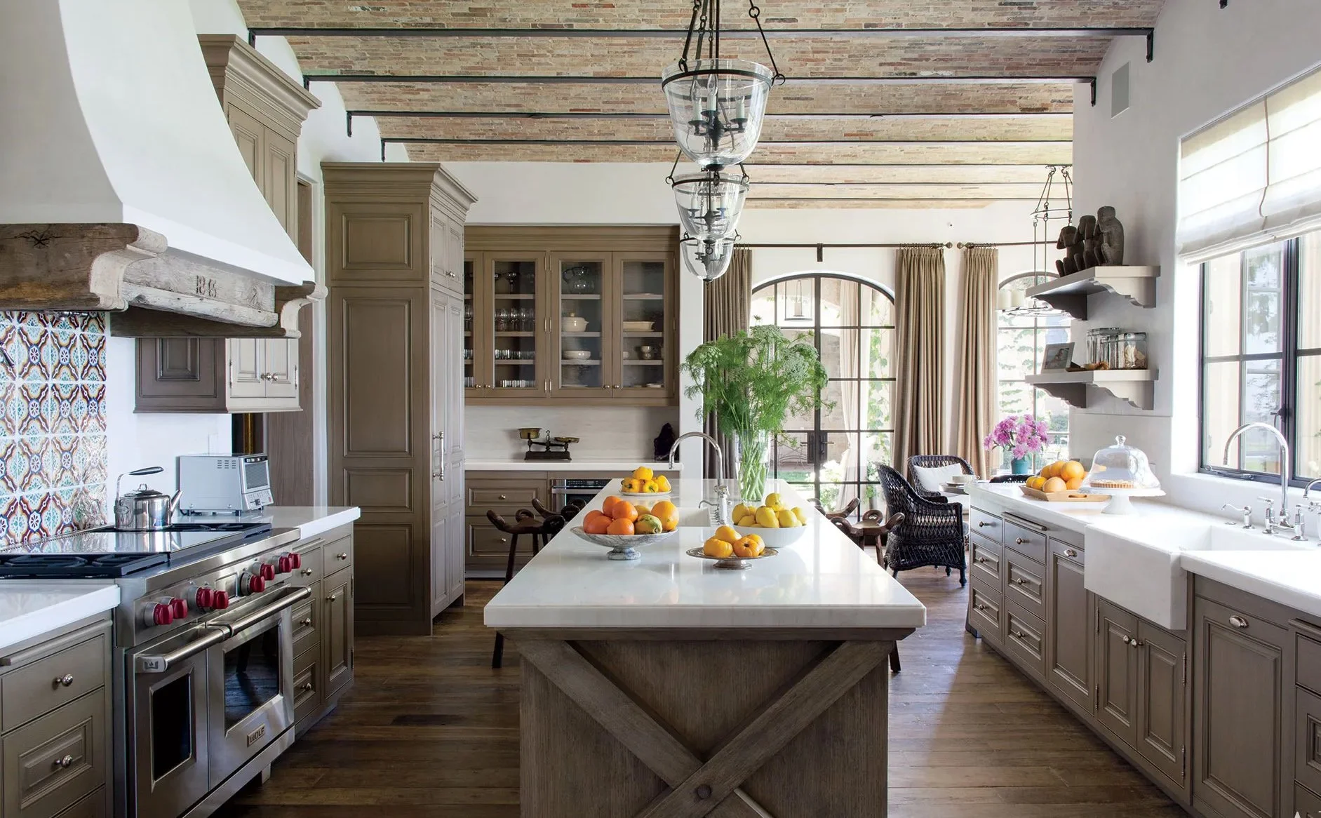 A spacious kitchen with a central island topped with bowls of fruit, beige cabinetry, a large window with beige curtains, and decorated with plants and decorative items.