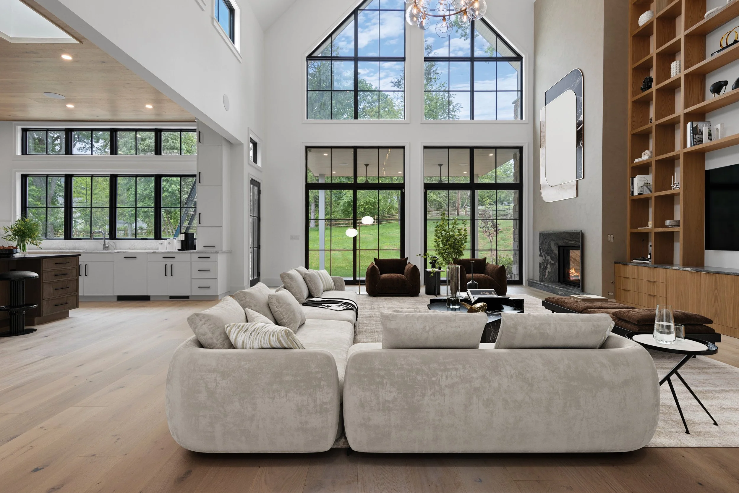Modern living room with large windows, high vaulted ceiling, and neutral-colored furniture. There's a white sectional sofa, a black coffee table, a fireplace, and a built-in wooden bookshelf.