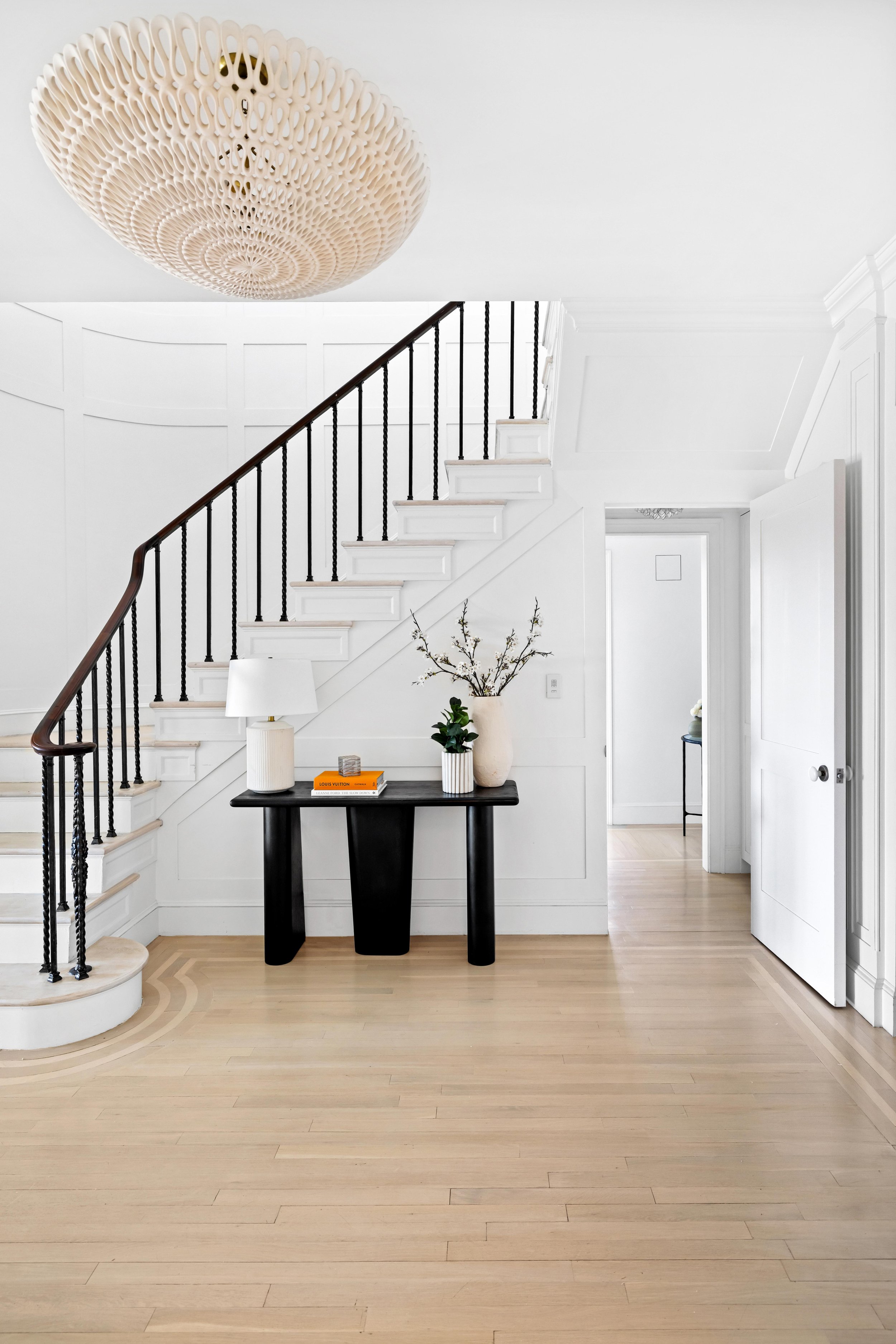 The image shows a minimalist interior space with a white staircase and walls, a black console table with decorative items including a white lamp, books, and a vase with branches and green leaves. A large, intricate hanging light fixture is mounted on