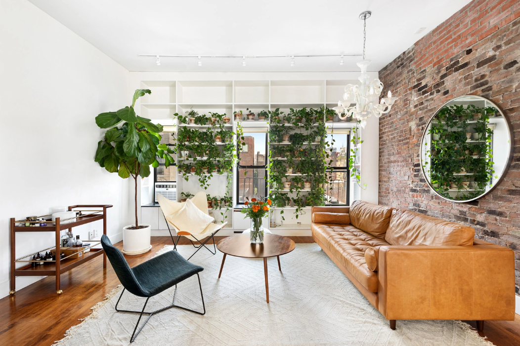 Living room with a brick accent wall, leather sofa, wooden coffee table with flowers, black and white chairs, large wall of plants, chandelier, and round mirror.