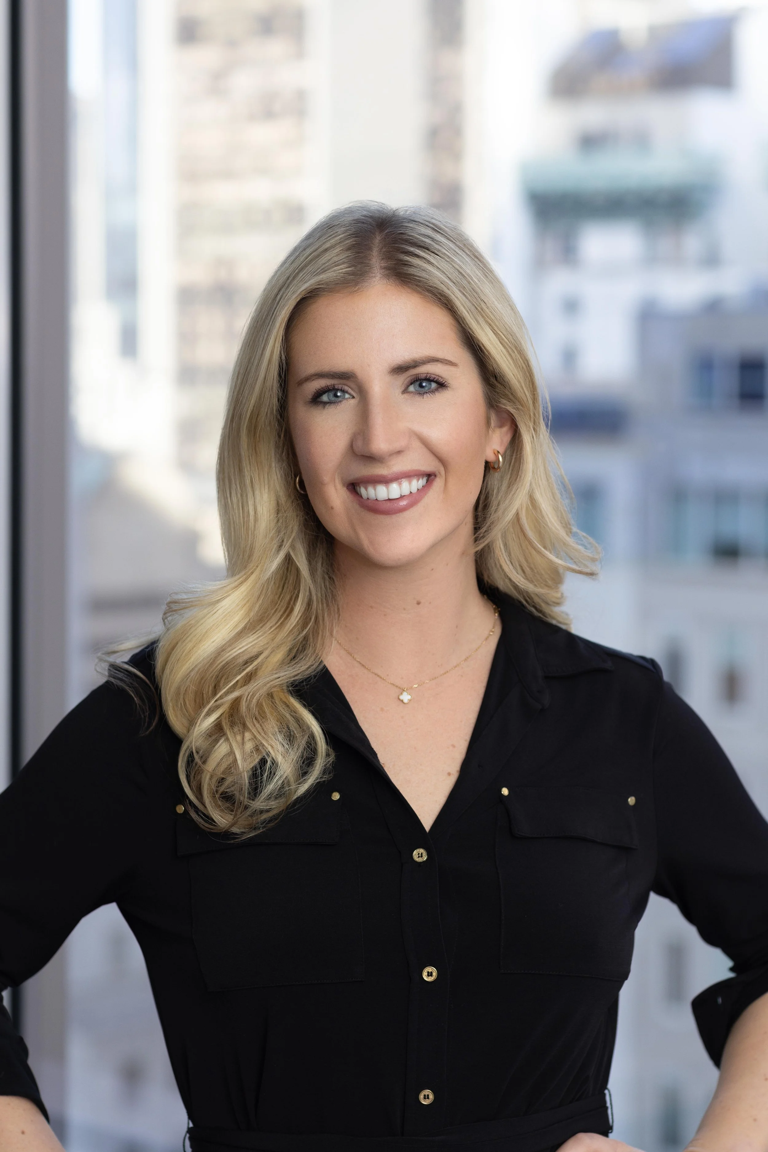 A smiling woman with blonde hair, wearing a black button-up shirt and jewelry, standing in front of a window with city buildings outside.