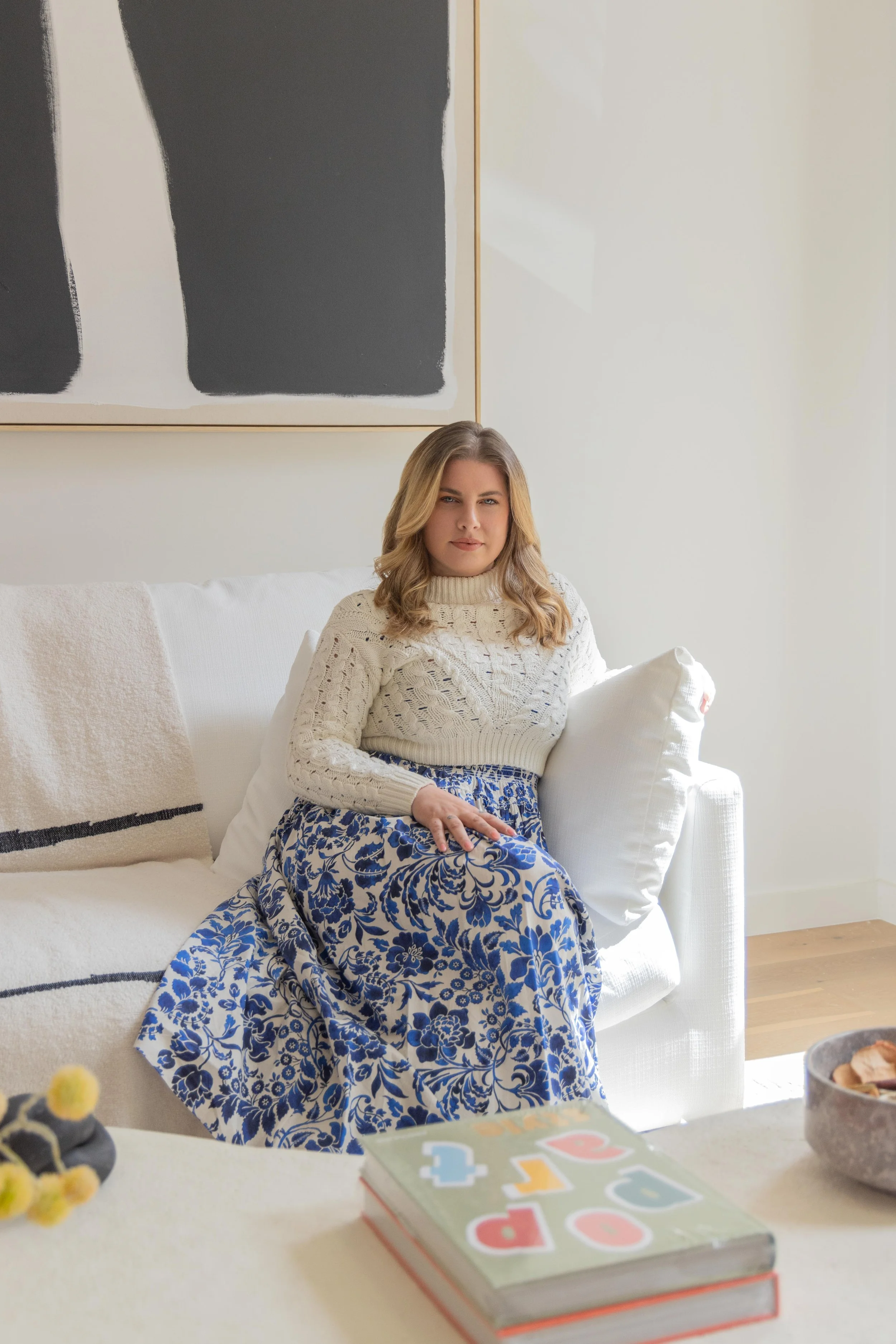 A woman with blonde hair sitting on a white sofa in a bright living room, wearing a cream-colored sweater and a blue and white floral skirt, with abstract black and white artwork on the wall behind her.