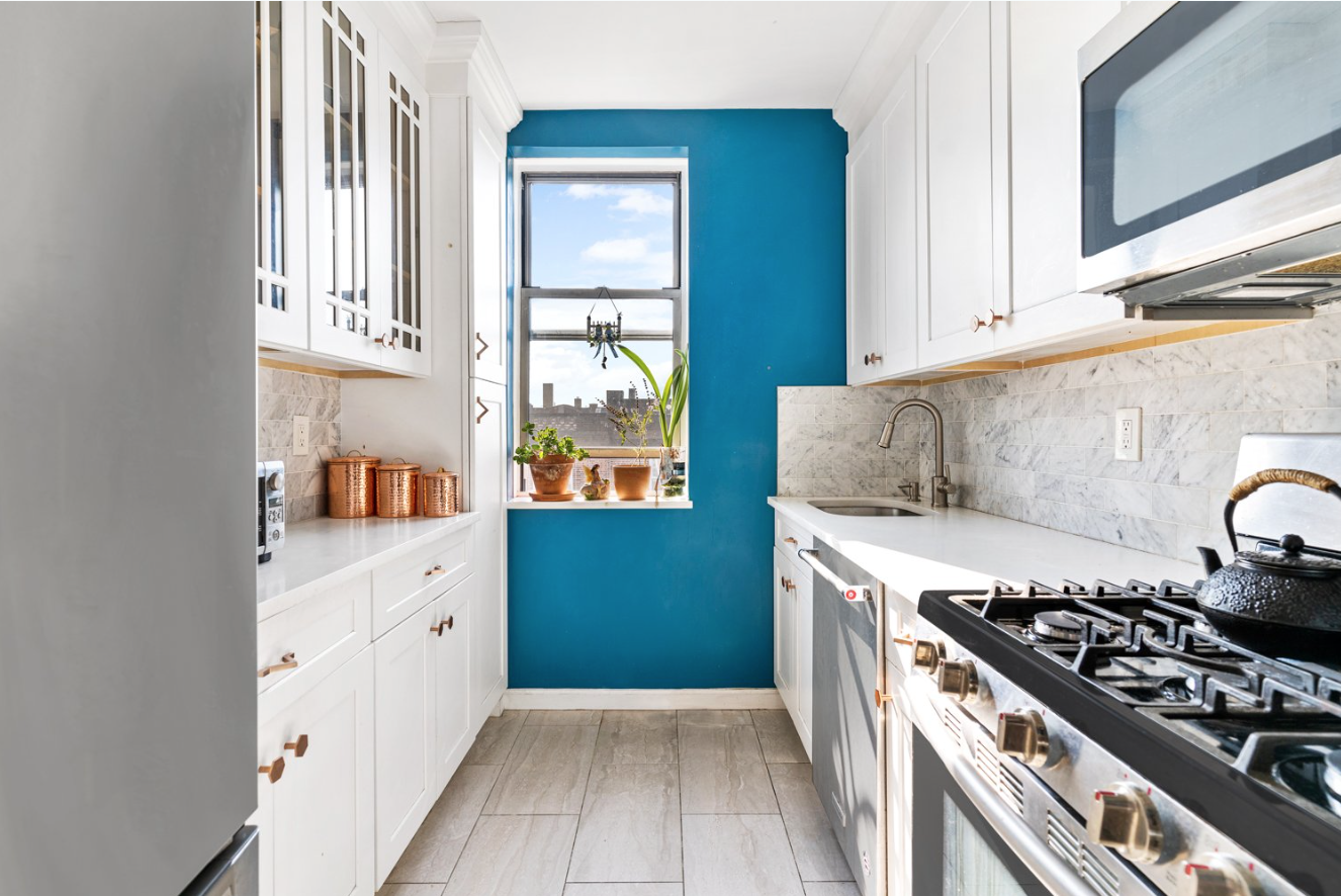 Bright kitchen with white cabinets, a blue accent wall, marble backsplash, and a window with potted plants, a black teapot on a stainless steel stove.