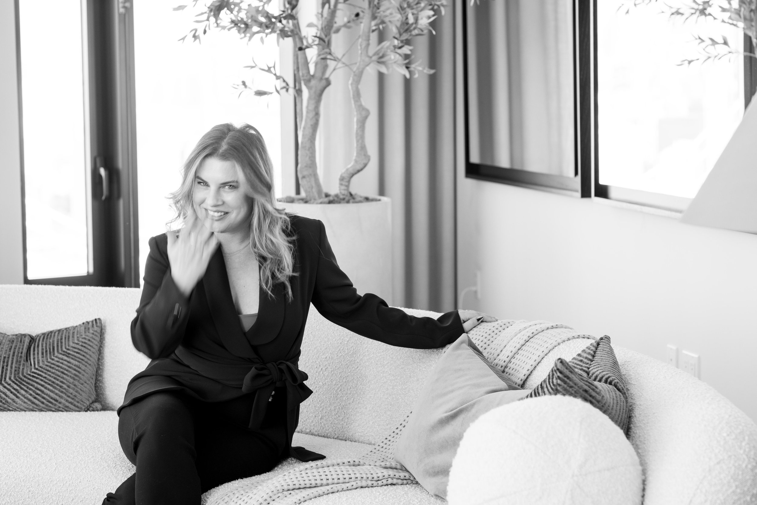 A woman with wavy hair smiling and sitting on a couch in a bright, modern living room, with a potted plant and windows in the background.