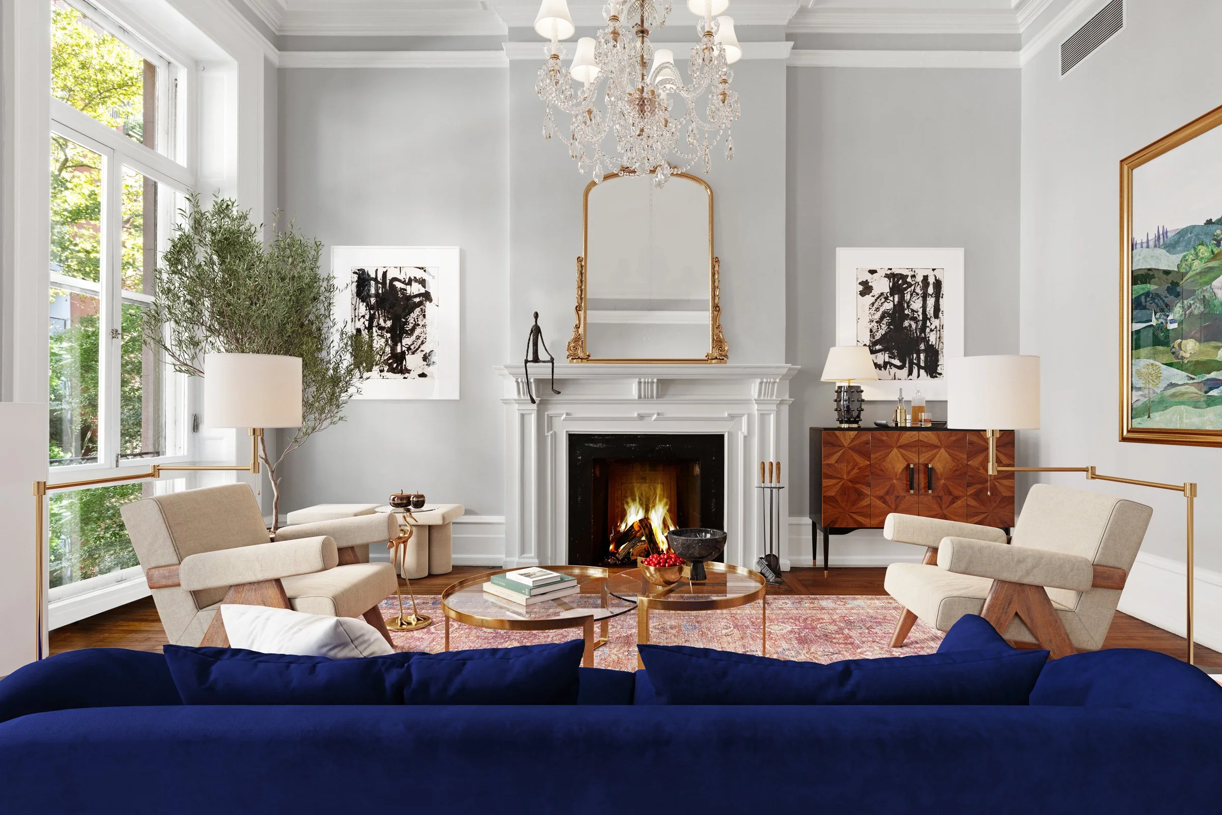 Living room with a lit fireplace, two beige armchairs, a navy sofa, a large window with greenery outside, a chandelier, artwork on the walls, a wooden sideboard, a mirror above the fireplace, and a coffee table with books and decorations.
