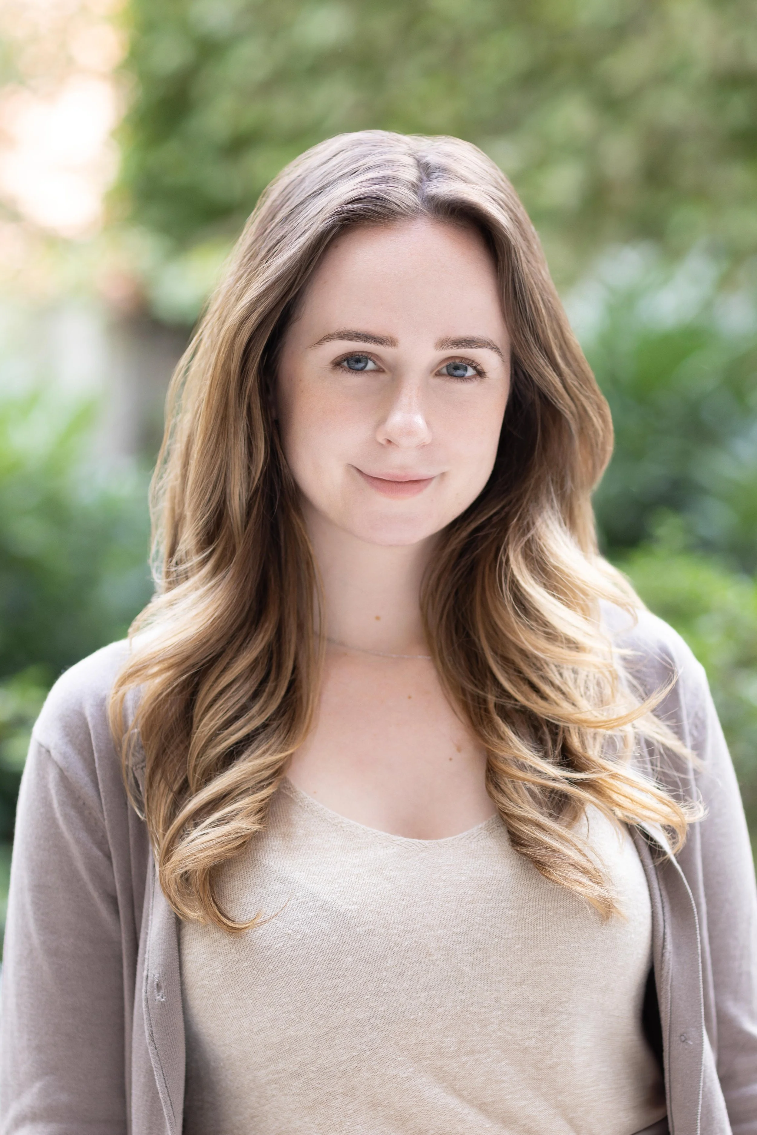 A young woman with long, wavy brown hair and blue eyes smiling softly outdoors, wearing a beige top and a light gray jacket, with a blurred green background.