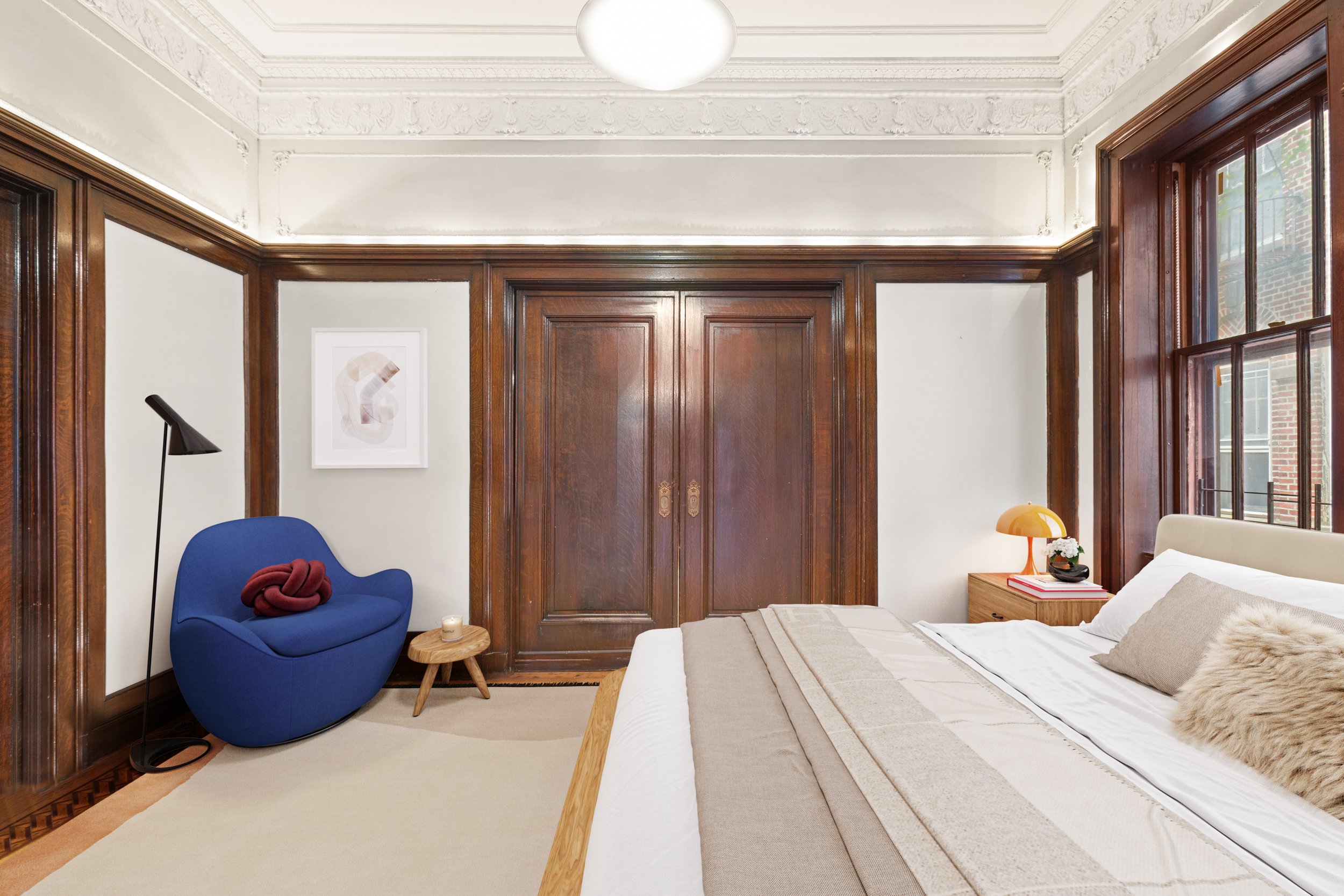 A bedroom with a wooden bed, nightstand with a lamp, a blue armchair, a small wooden side table, and large windows with wooden trim.